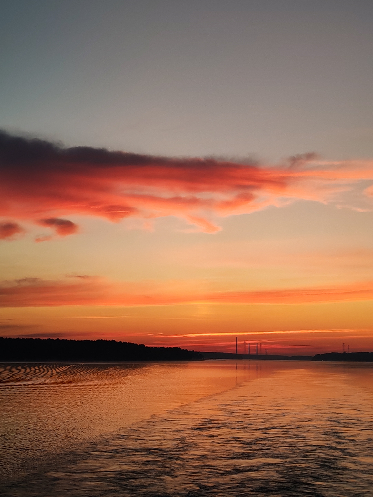 The image depicts a tranquil sunset over a body of water, likely a river or lake, near the city of Ploski, Russia. The sky transitions from a pale, muted blue at the top to vibrant hues of pink, orange, and red as it approaches the horizon. Wispy clouds, illuminated by the setting sun, are scattered across the sky, some tinged with deep purple.
A dark, silhouetted treeline borders the distant shore, suggesting a natural landscape. In the middle distance, several tall, industrial structures, possibly smokestacks or power transmission towers, are visible against the horizon, hinting at the proximity of human activity or infrastructure.
The water's surface is calm, with gentle ripples creating a textured pattern, reflecting the warm colors of the sky. A faint wake is visible on the water, indicating that a vessel may have recently passed through the scene. The overall atmosphere is serene and picturesque, capturing the natural beauty of the sunset.