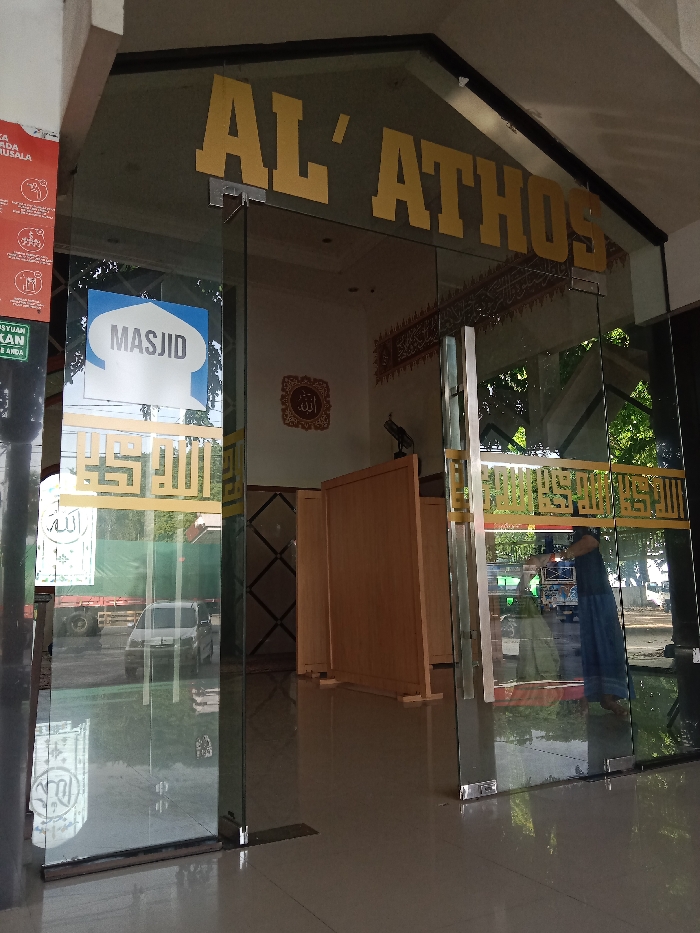 The image displays the glass entrance of a building identified as "AL'ATHOS" and a "MASJID" in Wonoketingal, Indonesia. Gold block letters spell "AL'ATHOS" above the transparent glass doors and sidelights. A blue and white sign reading "MASJID" in a mosque dome shape is affixed to the left glass panel, below which is gold Arabic calligraphy. A red informational poster is partially visible on the far left. Inside, the tiled floor is reflective. Multiple freestanding light brown wooden partitions are arranged. A dark object resembling a stylized bird or abstract sculpture rests on a dark pedestal behind the partitions. Calligraphy in Arabic script adorns the interior walls. A person wearing a long blue garment is partially visible on the far right, seen through the reflection on a glass door. Reflections on the glass also show parts of a white van, a red truck, trees, and an outdoor ground surface, indicating the building faces a street or parking area.