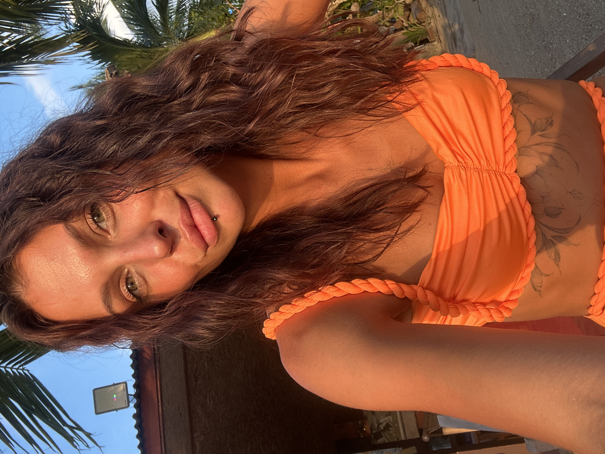 A woman with long, wavy brown hair and green eyes is taking a selfie outdoors. She is wearing an orange bikini with braided straps and ruffles. She has a lip ring and a tattoo on her torso. The background includes palm trees, a building with a light fixture, and a clear blue sky. The lighting suggests it is either early morning or late afternoon. This location appears to be in a tropical environment, consistent with Ko Samui District, Thailand.