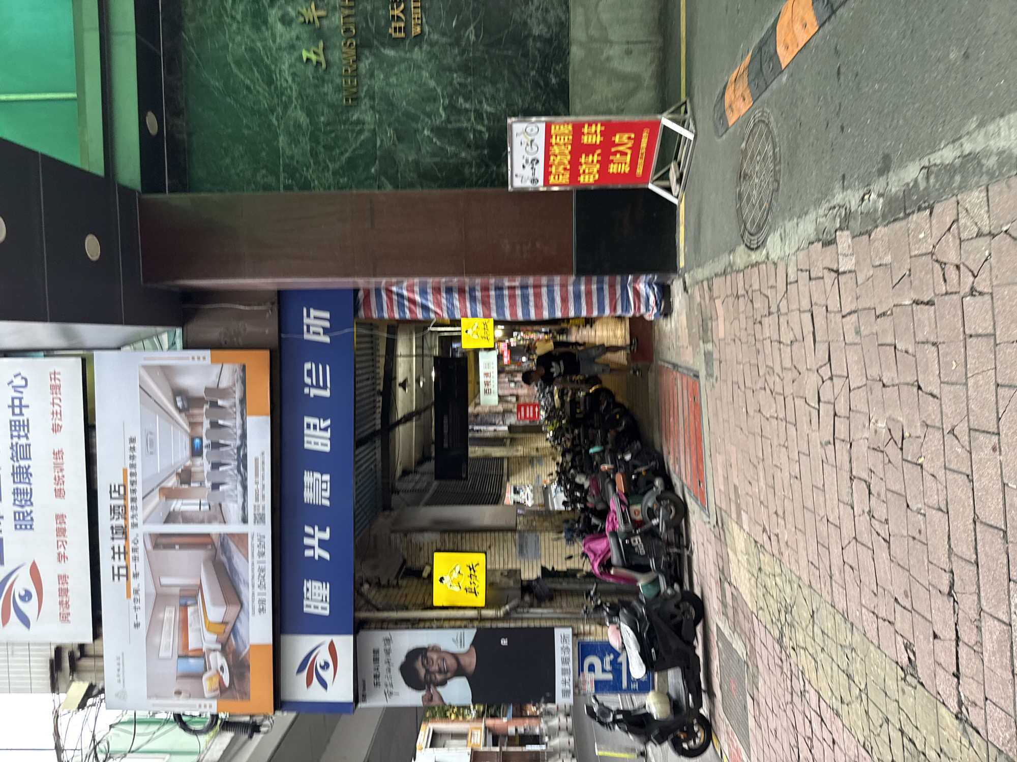 The image shows a street scene in Yuexiu District, China, featuring a row of parked motorcycles and scooters. In the background, several signs are visible, including one for "五羊城酒店" (Five Rams City Hotel) and another for an "eye clinic." A red and white striped banner hangs on a building. The ground is paved with red bricks, and a yellow line marks a path. A blue parking sign with a white 'P' is also visible. The overall environment appears to be an urban commercial area.
