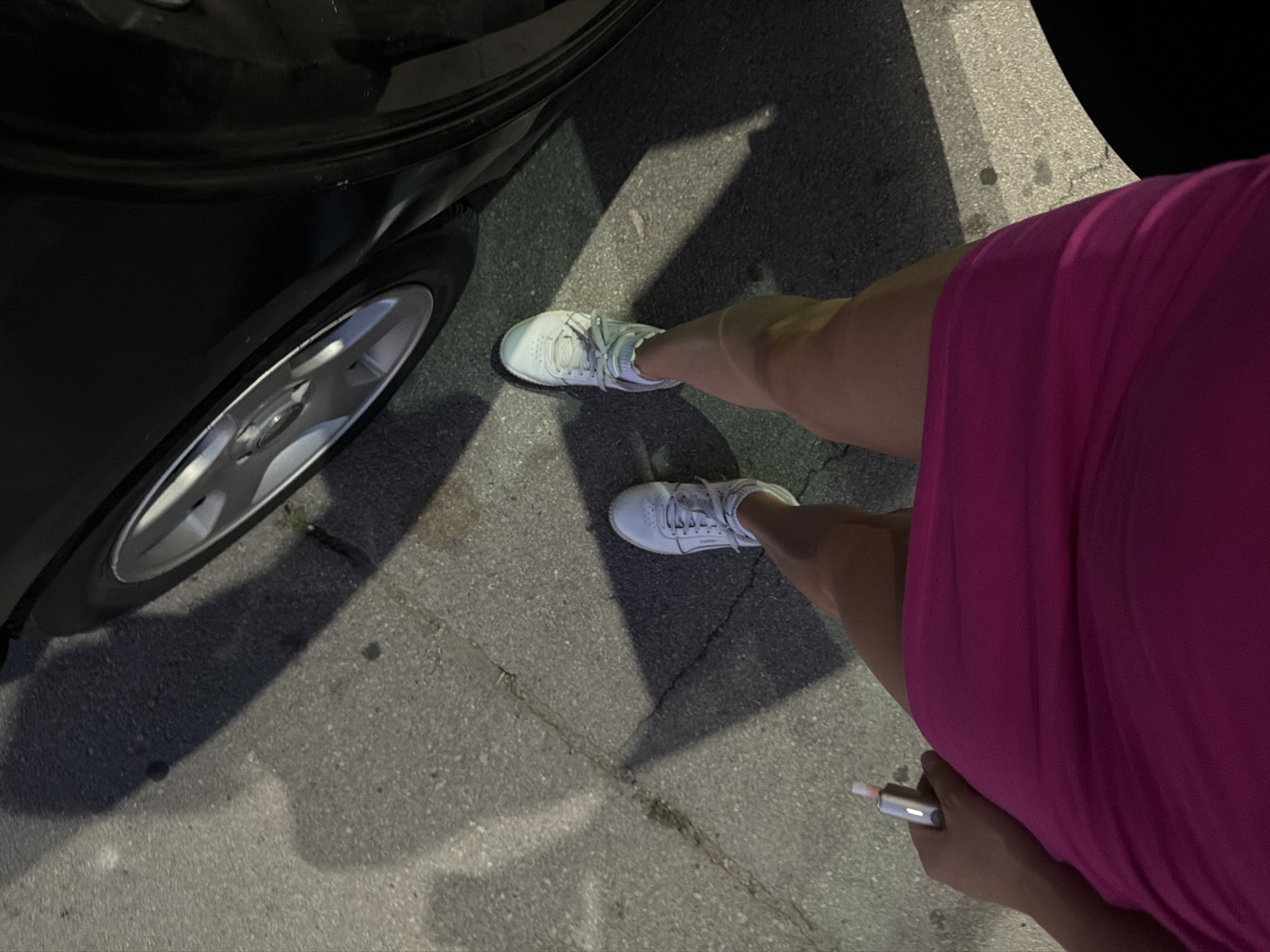 The image shows a first-person perspective of a person standing on a paved surface, possibly a sidewalk or street, next to a parked car. The person is wearing white sneakers and a bright pink dress. In their left hand, they are holding a silver object with a pink tip, which appears to be a vape pen. Shadows are cast on the ground from an external light source, suggesting it is daytime or evening. The environment is urban, with cracks visible in the pavement. The specific location in the city of Orhei, Moldova, is not discernible from the visual information provided.