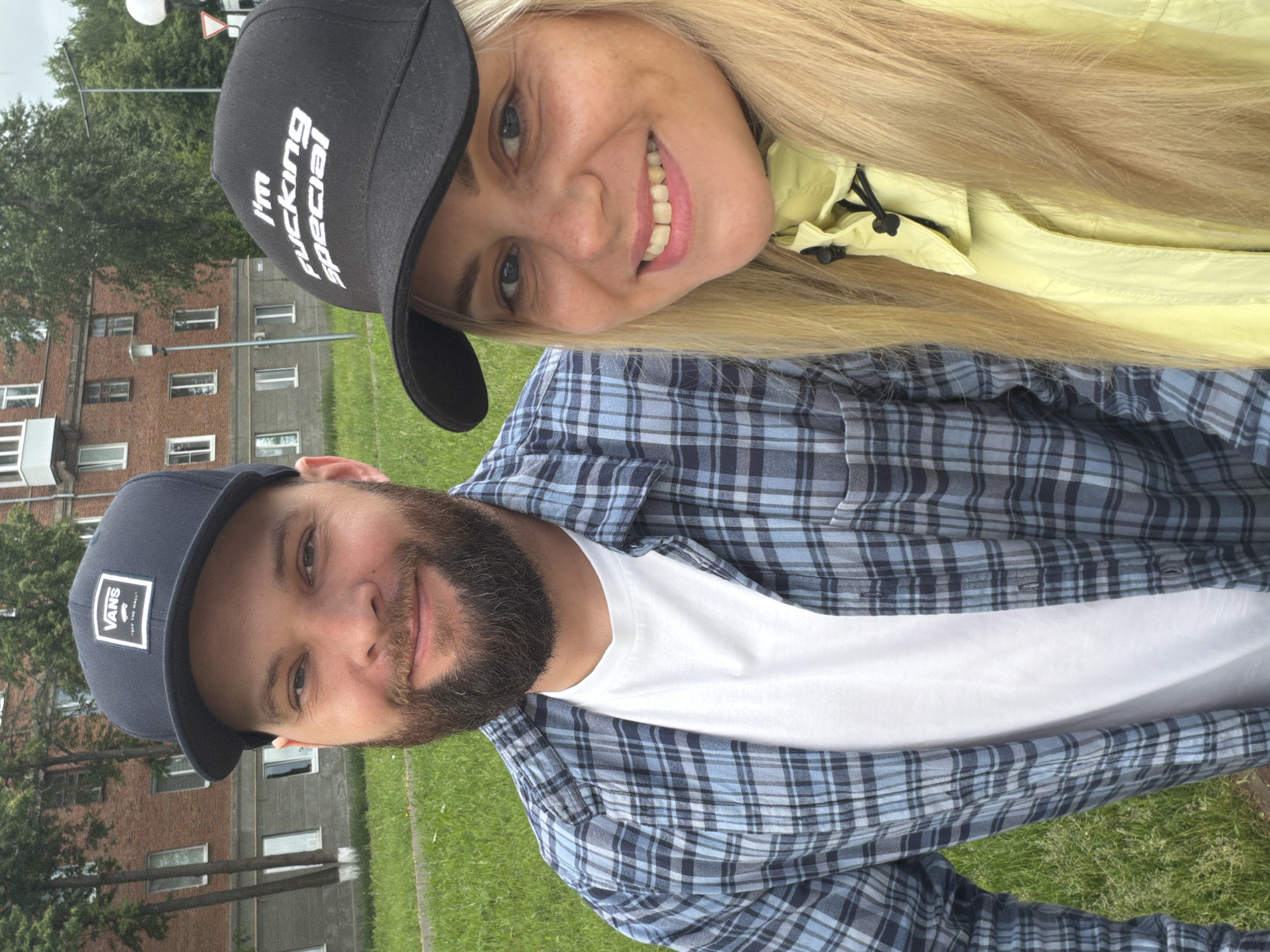 The image depicts two individuals, one male and one female, posing for a photograph outdoors in what is identified as the city of Votkinsk, Russia. The male subject, positioned on the left, has a light skin tone, dark eyes, and a full dark beard. He wears a black baseball cap with a white "VANS OFF THE WALL" logo, an open blue and grey plaid long-sleeve shirt, and a white crew-neck t-shirt. He is smiling, revealing his teeth. The female subject, positioned on the right, has a light skin tone, light blue eyes, and visible freckles on her cheeks. She has long blonde hair and wears a black baseball cap with the white text "I'm fucking special". She also wears a bright yellow jacket with a high collar and black toggle details. She is smiling, revealing her teeth, with a noticeable gap between her two upper front incisors. Both subjects are looking directly at the camera. The background features a multi-story brick building, partially obscured by green trees. A patch of green grass is visible in the foreground, along with a paved path or sidewalk. A grey lamppost and a street sign are visible on the right side of the background. The sky appears overcast.