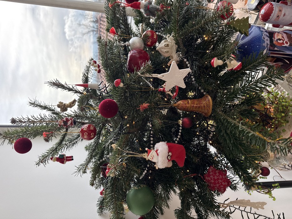 An indoors scene in Rousset, France, depicts a densely decorated Christmas tree positioned in front of a large window. The tree is adorned with various ornaments, including multiple red velvet spheres, red "HO HO HO" labeled spheres, a large green sphere, a silver sphere, and a red apple-shaped ornament. Figures such as several small Santas (one in a red stocking, another holding a gift), a golden angel, a white porcelain-like angel, and a grey-bearded gnome with a red hat are distributed among the branches. Additional decorations include white star-shaped ornaments, a gold-toned bell, red mushroom miniatures, a white bead garland, and numerous warm white fairy lights. A red star ornament with a holly pattern and a white lace-like decoration are also present. The base of the tree shows glimpses of a small green plant with berries. Through the window, an overcast sky is visible above distant bare trees and several buildings featuring characteristic red-tiled roofs, indicative of the surrounding Rousset landscape. The lower right edge of the frame reveals parts of additional festive objects, including a blue container with white stars and a red Santa figurine.
