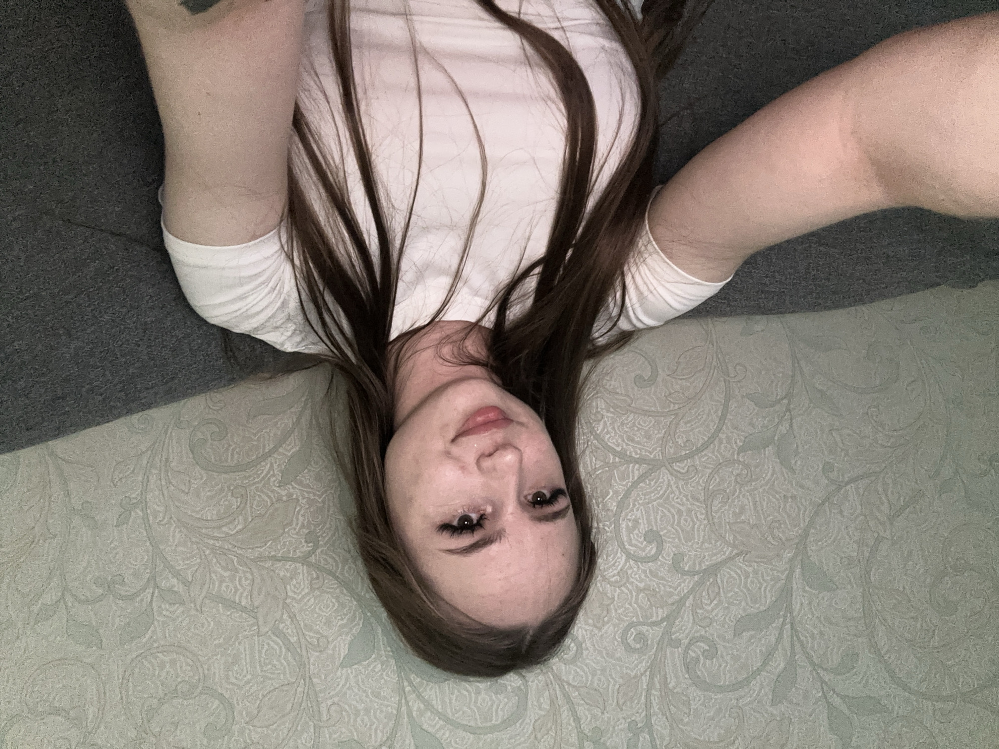 A young woman with long, brown hair and long eyelashes is seated on a grey couch. She is wearing a white t-shirt. The wall behind her is decorated with a light green, floral wallpaper. The image appears to be a selfie taken from a slightly low angle. The woman is looking directly at the camera with a slight smile. Her left arm is extended, suggesting she is holding the camera.