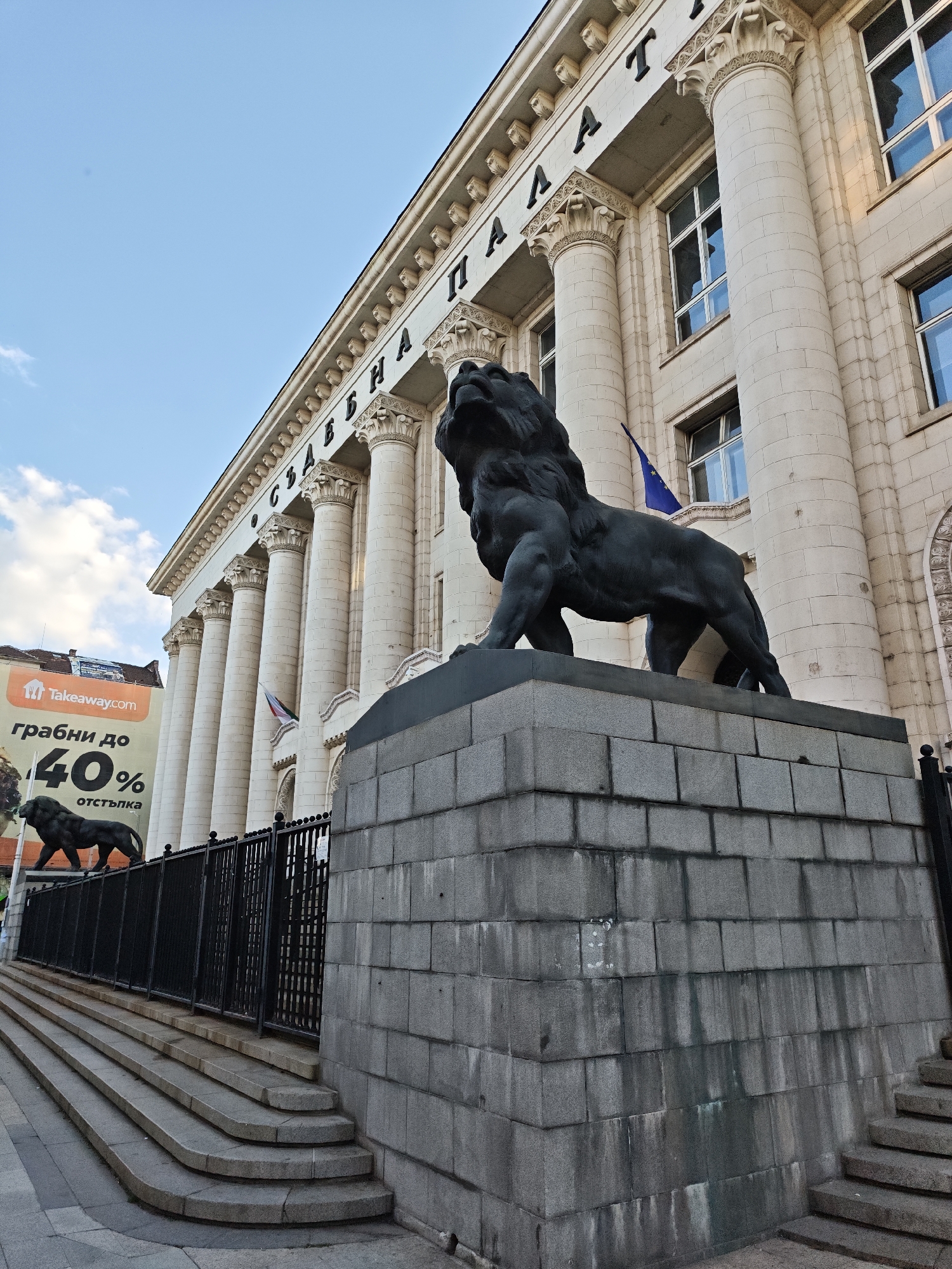 The image shows the Palace of Justice in Sofia, Bulgaria, with a prominent lion statue in the foreground. The statue is black and stands on a grey stone pedestal. A second, similar lion statue can be seen behind a black iron fence on the left. In the background, the Palace of Justice building is characterized by its white stone facade, classical columns, and a sign in Cyrillic script above the columns. To the left, an advertisement for "Takeaway.com" is visible with text and a lion image. The sky is blue with a few clouds. The steps leading up to the fence are made of grey stone. No people are visible in the image.