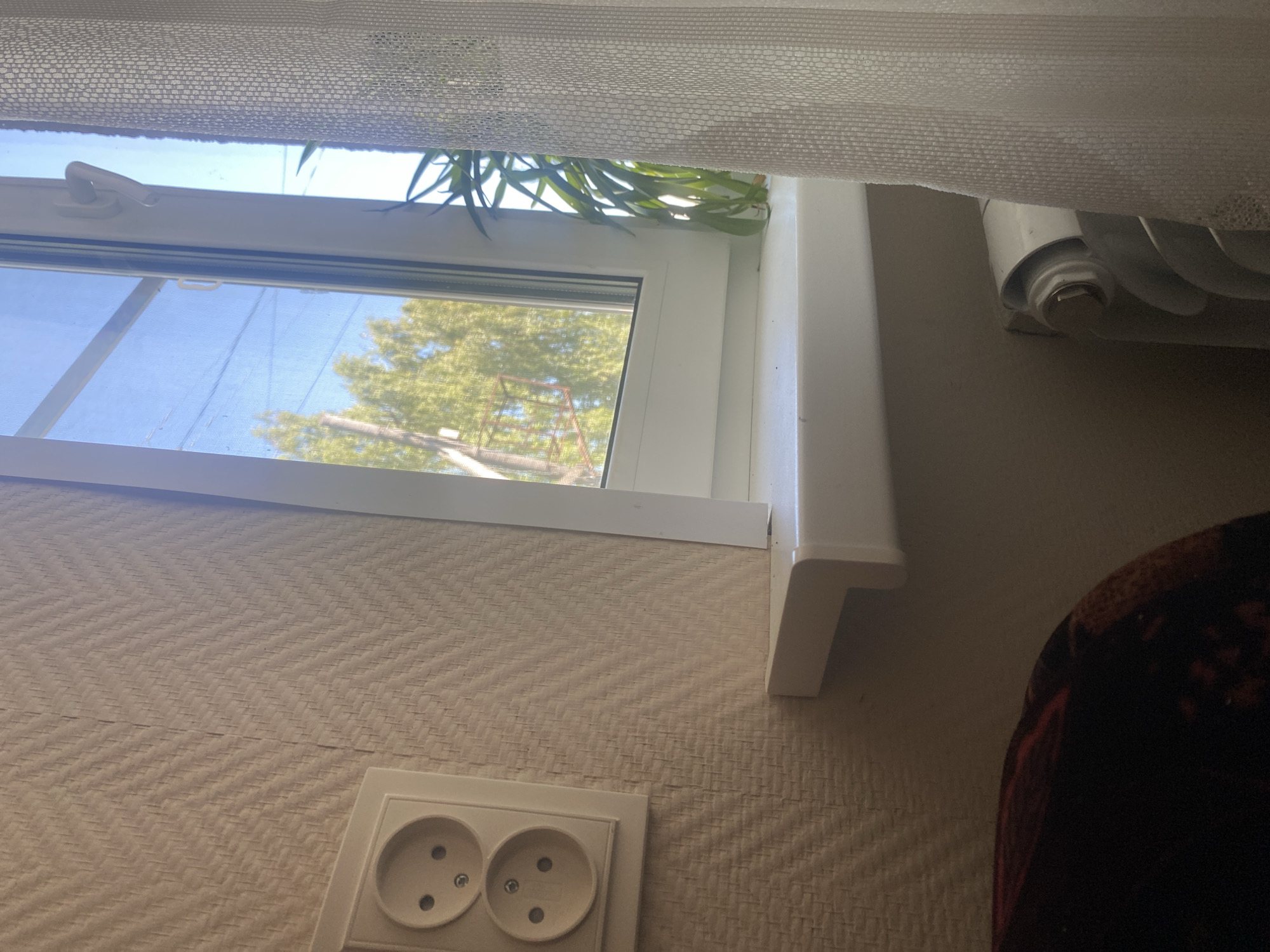 This image shows an interior view looking out a window. The window is partially covered by a sheer white curtain. Through the window, green trees and a blue sky are visible, suggesting it is daytime. A white windowsill runs along the bottom of the window. To the right of the window, a white radiator with a curved design is partially visible. Below the window, on a wall with textured wallpaper, is a double electrical outlet with two sockets. To the far right of the image, a portion of a dark, patterned blanket or piece of furniture is visible. The scene appears to be in a domestic setting, likely a room in a house or apartment in Afanasyevo, Russia. There is no discernible activity taking place, and the overall mood is neutral and ordinary. No text is visible in the image.