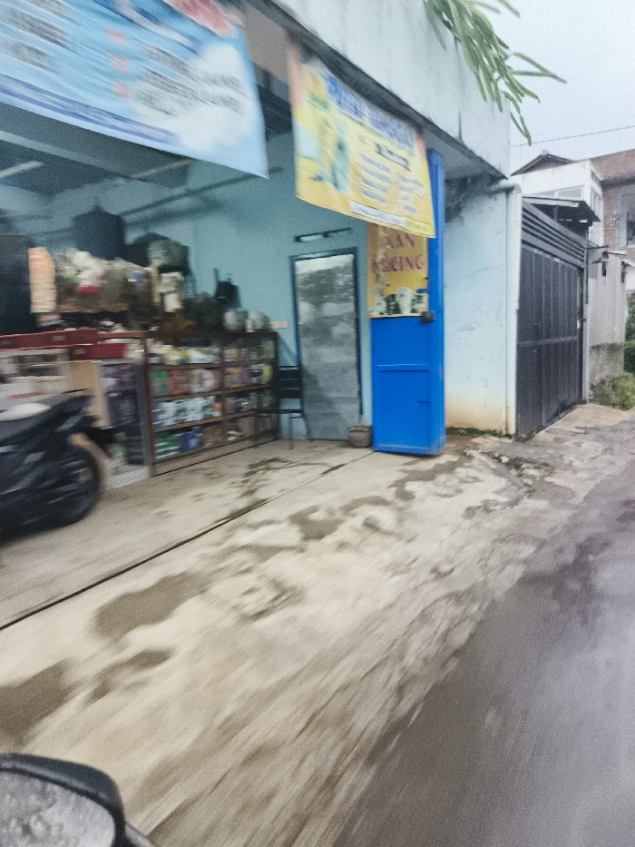 The image shows a view from the street, looking towards the front of a shop in Cimenyan, Indonesia. The shop has a blue door with a yellow sign that says "Ban Racing" in white lettering. The shop also has a black gate to the right, and a window with a blue sign partially visible. A motorcycle is parked to the left of the shop, near the front, with its front tire visible. There are shelves full of items in the shop, and a chair is visible on the right. The street is wet, likely from rain. The environment is urban and there is a building in the background.