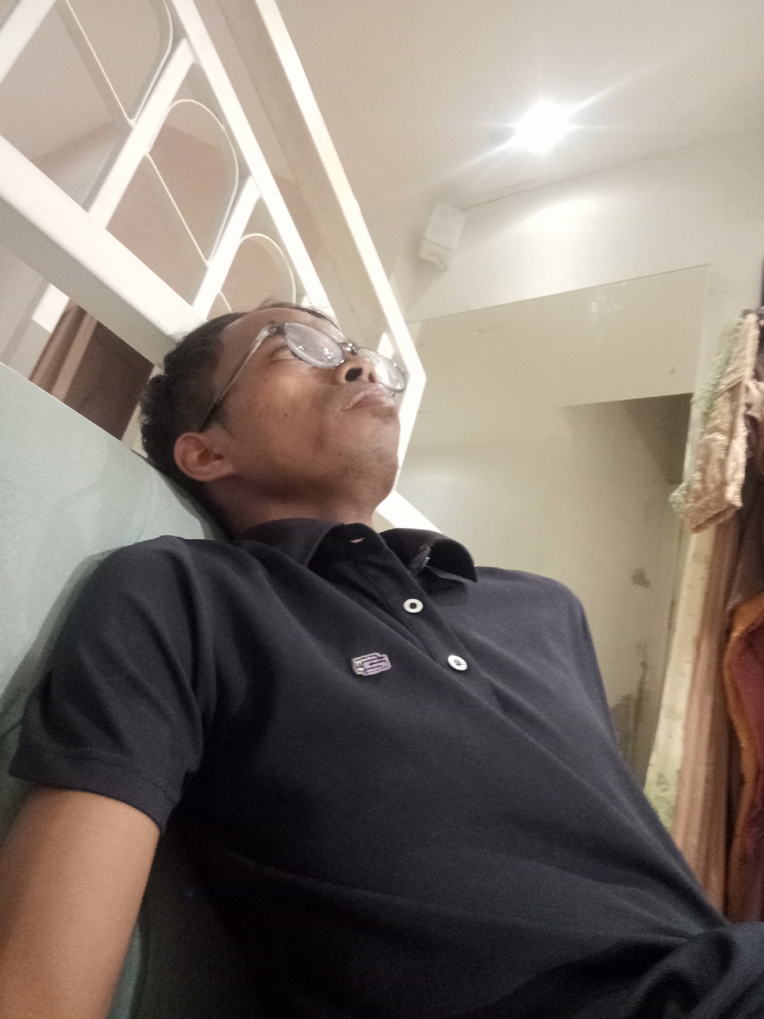 A medium-shot, low-angle, interior photograph shows a man wearing glasses and a black polo shirt, looking up and to the right with his head tilted back. The man has dark skin and short, dark hair. He is seated next to a light-colored upholstered surface, possibly a couch. Behind him, a white railing with a decorative pattern runs diagonally from the bottom left to the top left of the frame, suggesting he is on a staircase or elevated platform. The ceiling is white and has a bright light fixture that creates a starburst effect. To the right of the man, there appears to be a wall and some draped fabric, possibly clothing or curtains. The scene is indoors and appears to be within a residential or public building. The photograph was taken in Larangan, Indonesia.