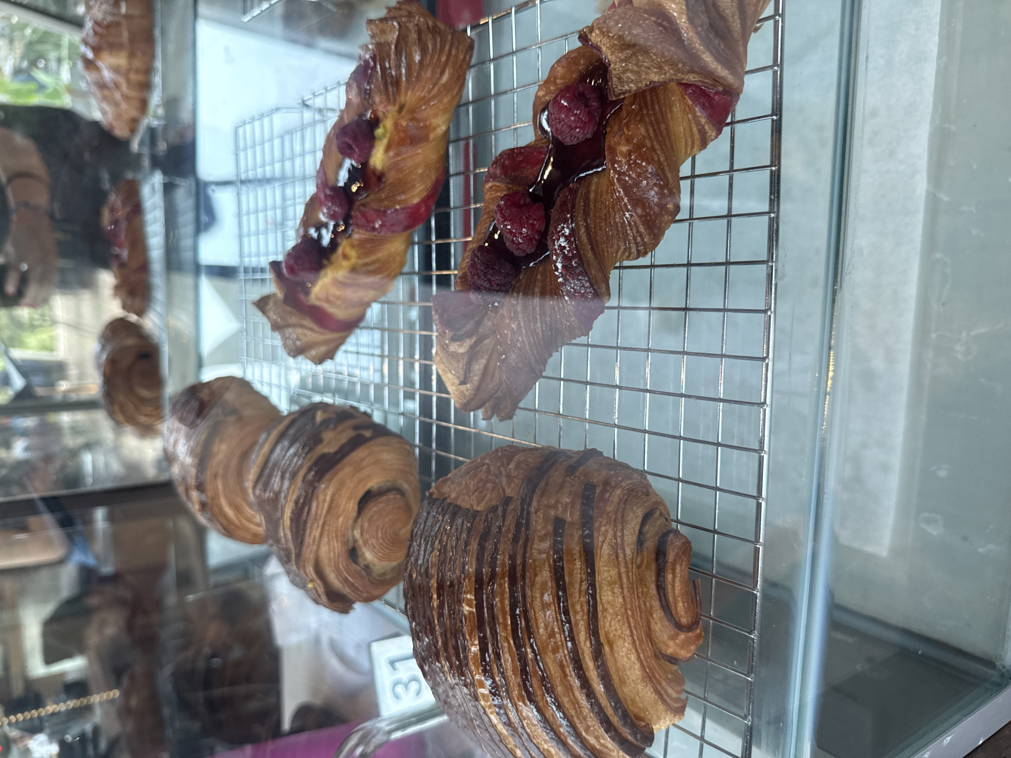 A display case holds various pastries. At the top are two twists with raspberries. Below them are three chocolate croissants. The location is San Pedro Garza García, Mexico.