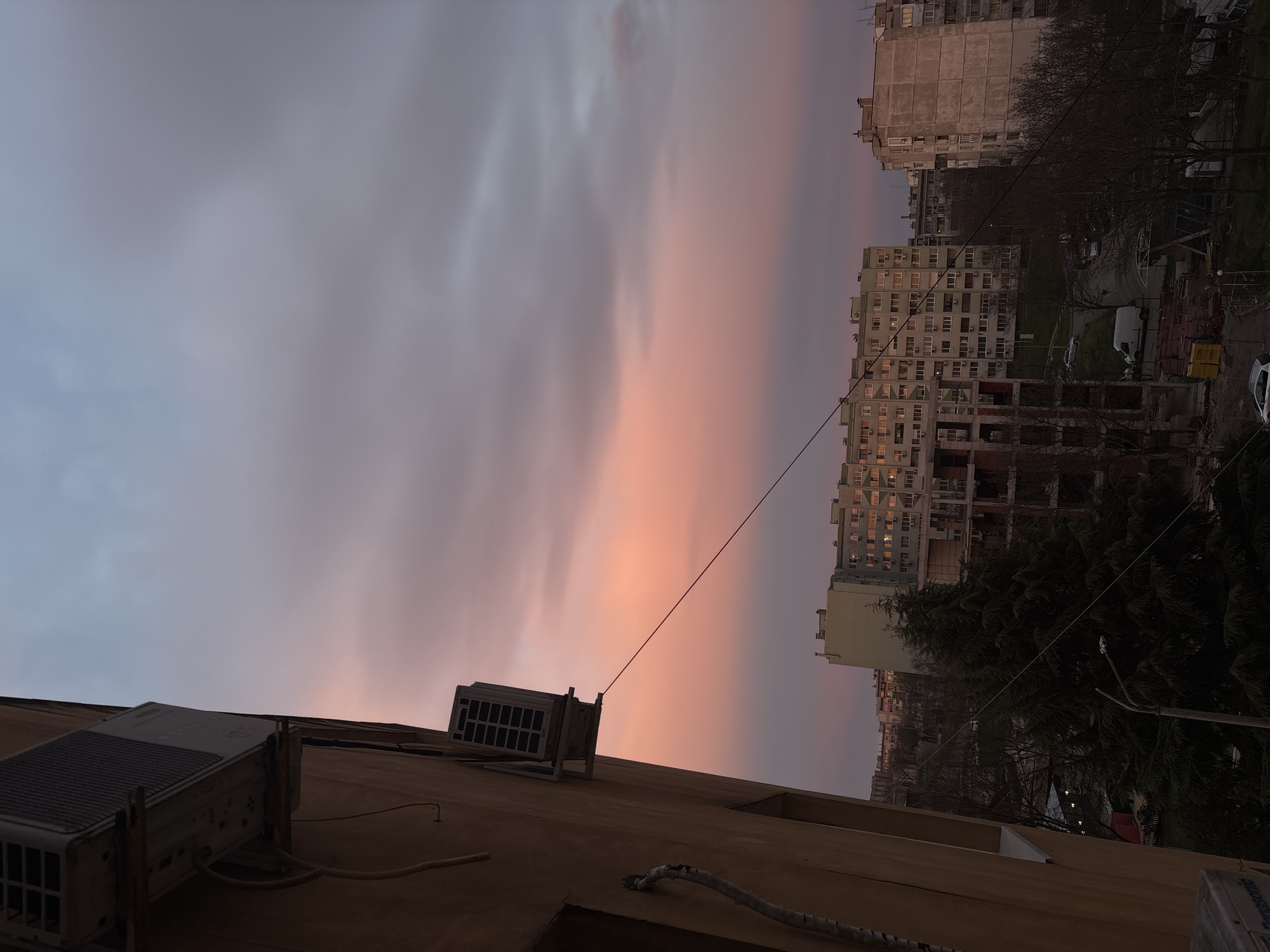 The image shows a sunset in an urban environment, possibly Burgas, Bulgaria. The sky is a blend of grey and pink hues. In the foreground, there's a building with air conditioning units attached to the exterior wall. The background reveals apartment buildings and trees. There are some cars parked in the street between the buildings.