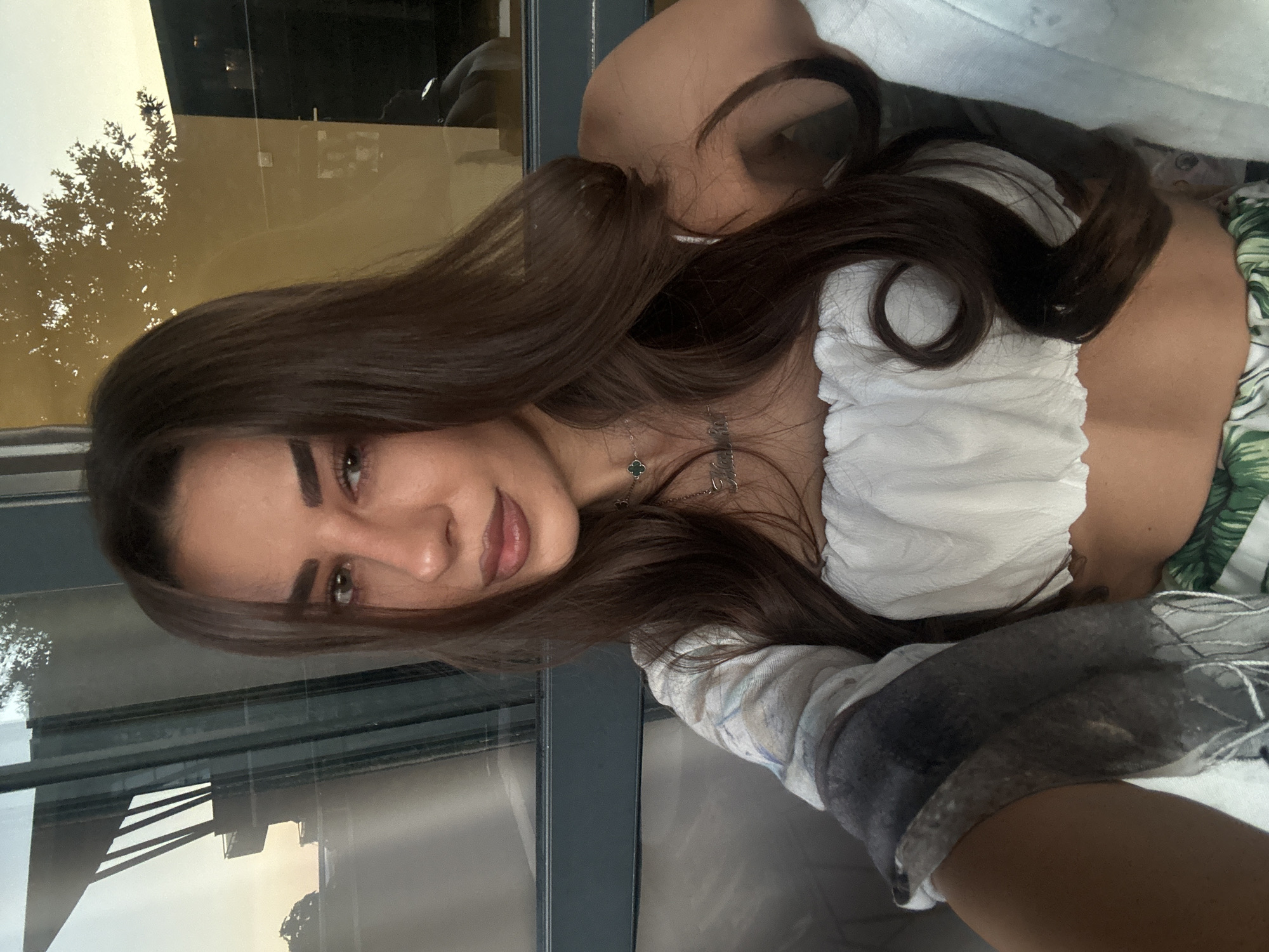 A young woman with long, dark brown hair is taking a selfie in front of a large window. She is wearing a white, ruffled tube top and a printed skirt with green leaves. A delicate silver necklace with a clover pendant and a nameplate is visible around her neck. The window reflects the golden hour sky and the silhouette of a building with architectural elements, possibly a modern structure. The background also shows the reflection of trees and some interior lighting. The woman is seated, as indicated by the slight upward angle of the camera and the way her arm is positioned to take the photo. The lighting suggests it is late afternoon or early evening. The scene is likely set at a restaurant or cafe with a view, possibly located in Nesebar, Bulgaria, given the architectural style in the reflection which could be indicative of contemporary structures found in such a location.