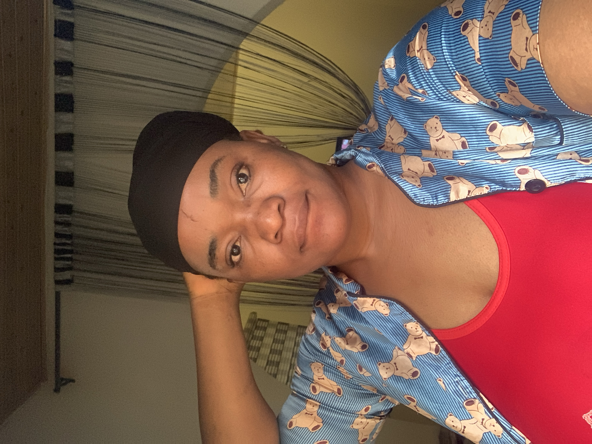 This image captures an adult woman, likely located indoors in Barakin Akawo, Nigeria. She is looking directly at the camera with a calm, friendly expression and a slight smile. Her head is tilted, and her left hand is visible, gently resting against her jaw or head, suggesting a relaxed posture.
The woman has a dark complexion and is wearing a black head covering. She is dressed in a vibrant red top, possibly a tank top or t-shirt, which is partially covered by an open, light blue shirt. The blue shirt features a pattern of thin, horizontal stripes and numerous light brown cartoon teddy bears. Some of the teddy bears are adorned with black bow ties.
The background indicates an indoor setting. To the left of the frame, a segment of what appears to be a wooden door frame or furniture can be seen. Behind her, there is a wall and a textured element resembling a window blind or a curtain with horizontal lines or slats. A portion of the wall near her head exhibits a yellowish hue. The lighting in the image is soft, implying artificial illumination or a naturally subdued indoor light. There is no visible text present in the image.
