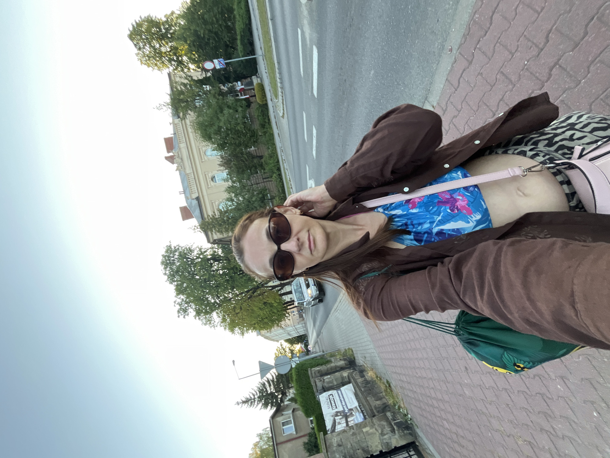 A woman with long brown hair and sunglasses is taking a selfie on a paved sidewalk next to a road. She is wearing a brown open-buttoned shirt over a blue and pink floral crop top, and zebra-print pants. She has a green drawstring bag hanging from her left shoulder and a pink crossbody bag. In the background, there is a street with a car and buildings, and trees lining the road. The sky is clear and blue.