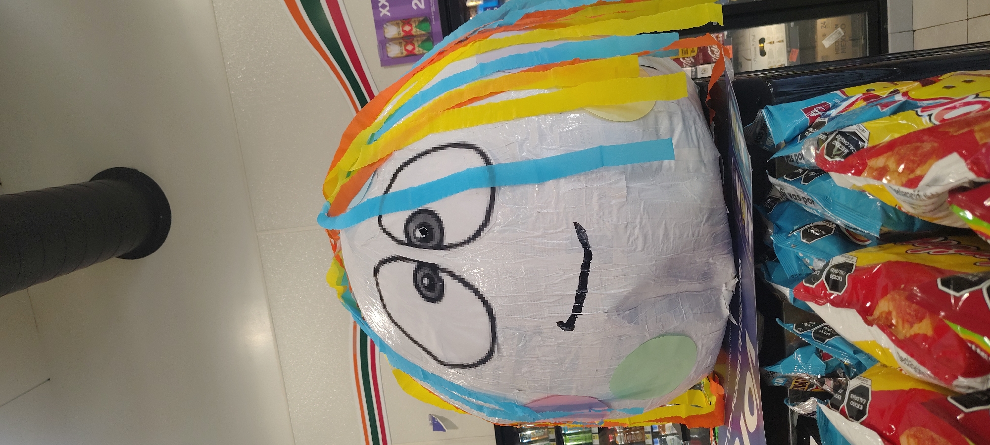 The image shows a piñata with a crude cartoon face and colorful crepe paper fringe on display for sale. The piñata is located in a 7-Eleven convenience store in San Miguel Acapantzingo, Mexico. It is situated next to bags of Sabritas brand potato chips. A ventilation duct is visible on the ceiling.