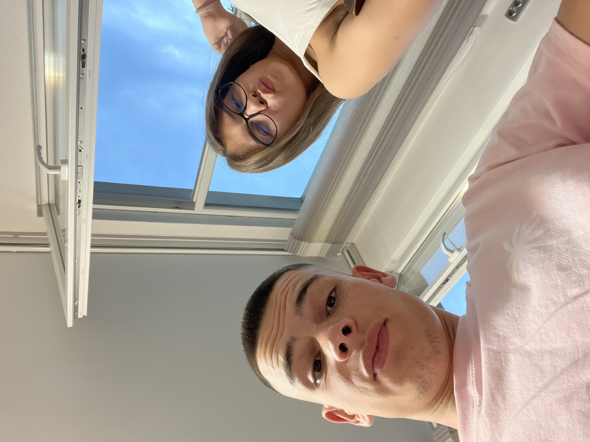 An upward-angle selfie-style photograph depicts two young individuals indoors, in Karavelovo, Bulgaria. In the left foreground, a male with a short buzz cut and light facial hair on his chin gazes directly at the camera, his lips slightly pursed. He wears a light pink t-shirt with a white palm tree graphic on the chest. In the upper right background, a female with long brown hair and black-rimmed glasses also looks towards the camera, her lips forming a pout. She wears a white sleeveless top. Behind the subjects, a white-framed window is open, revealing a clear blue sky. Vertical blinds or pleated shades are visible on the right side of the window frame.