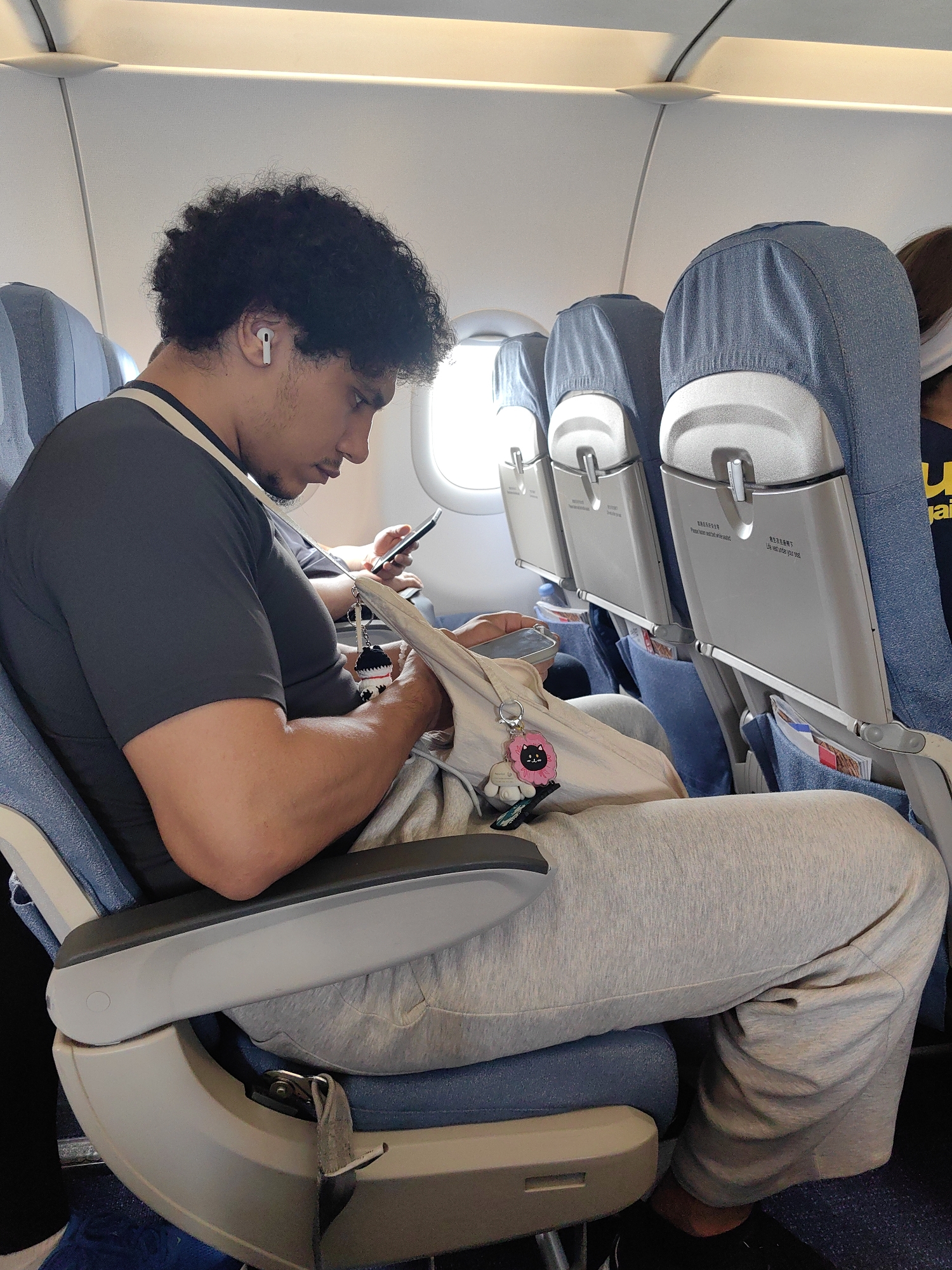 The image shows a person sitting on an airplane. The person is a man with dark, curly hair and an earbud in his left ear. He is wearing a dark gray t-shirt, a white shoulder strap, and light gray sweatpants. He is looking at a phone in his hand. There is a beige tote bag in his lap with a pink and black cat keychain attached.
The airplane seat is blue with a gray armrest. The seat in front of him is also blue, with a gray headrest cover and a gray tray table attached to the back. There is a safety instruction card in the seat pocket. The background shows other airplane seats and a window.