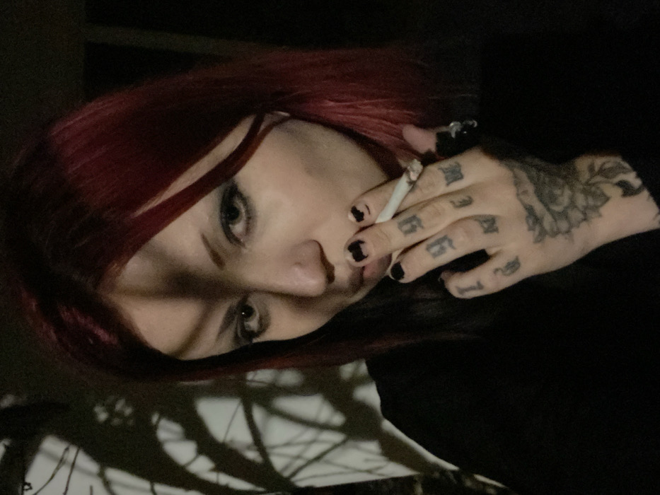 A close-up, chest-up portrait shot features a young adult female in Kunje, Montenegro. She has shoulder-length dark red hair, pale skin, and heavy dark eye makeup. A small silver ring is present in her left nostril. A distinct vertical shadow falls across the left side of her face. Her left hand is visible, holding a lit cigarette between the index and middle fingers, positioned near her mouth. Her fingernails are painted black, with some chipping evident. The fingers of her left hand display black script tattoos, and a larger black tattoo resembling a floral or mandala pattern covers the back of her hand. A black ring with an irregular shape is on her left ring finger. She wears a black long-sleeved garment. The background is dimly lit, with the shadows of bare tree branches cast onto a lighter surface on the left side of the frame, while the right side remains dark.