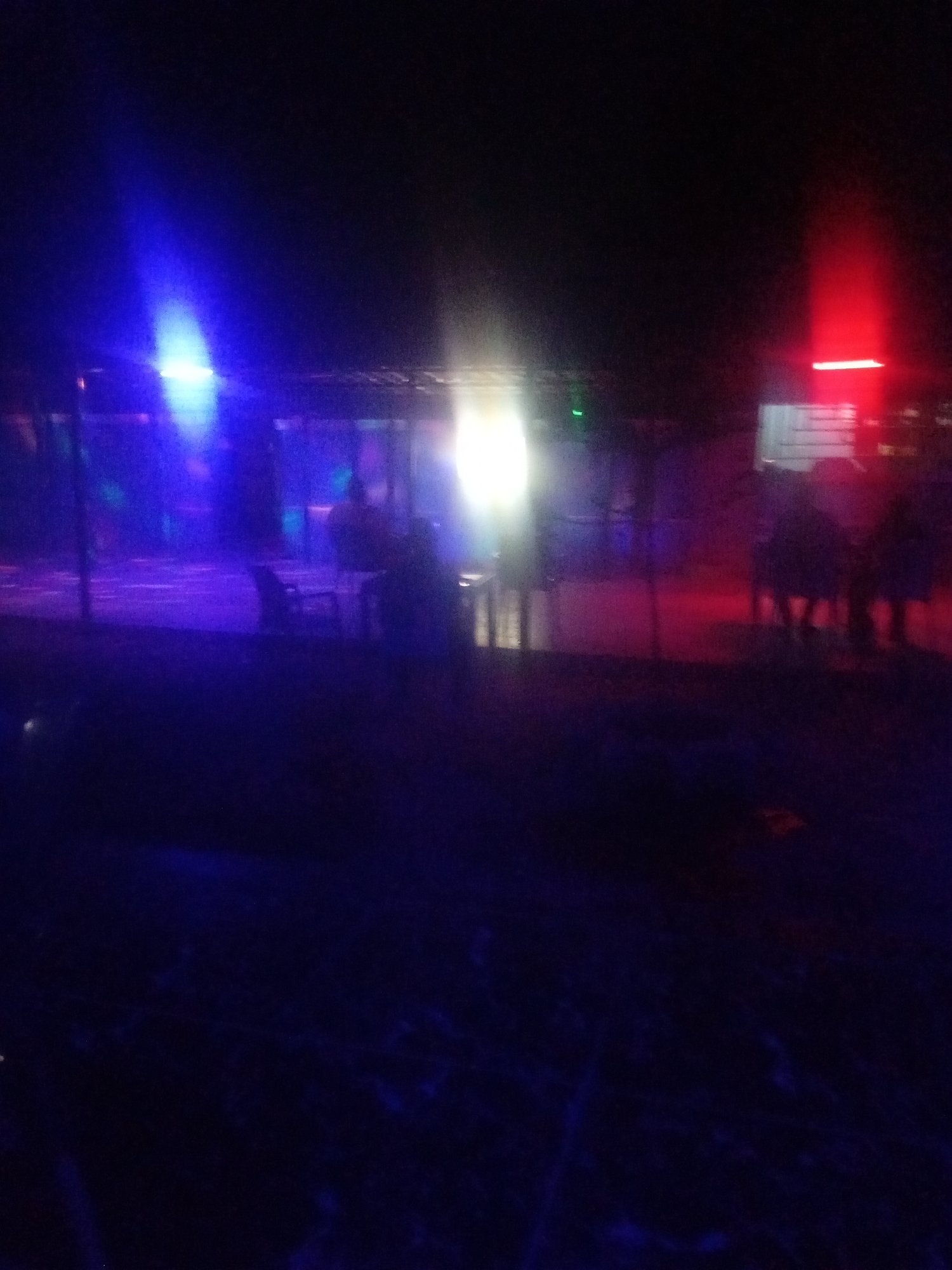 The image depicts an outdoor scene at night, likely an entertainment venue, illuminated by colored lights. In the foreground, a dark, unidentifiable area transitions to a mid-ground with a covered structure. This structure appears to have tables and chairs, suggesting a seating area. Beyond this, a fence or railing is visible. The background is largely dark, but features a bright, intense red light source on the right side and a bright blue light source on the left. Silhouetted figures are present within and around the covered structure, implying human activity. The overall atmosphere is suggestive of a party or gathering.