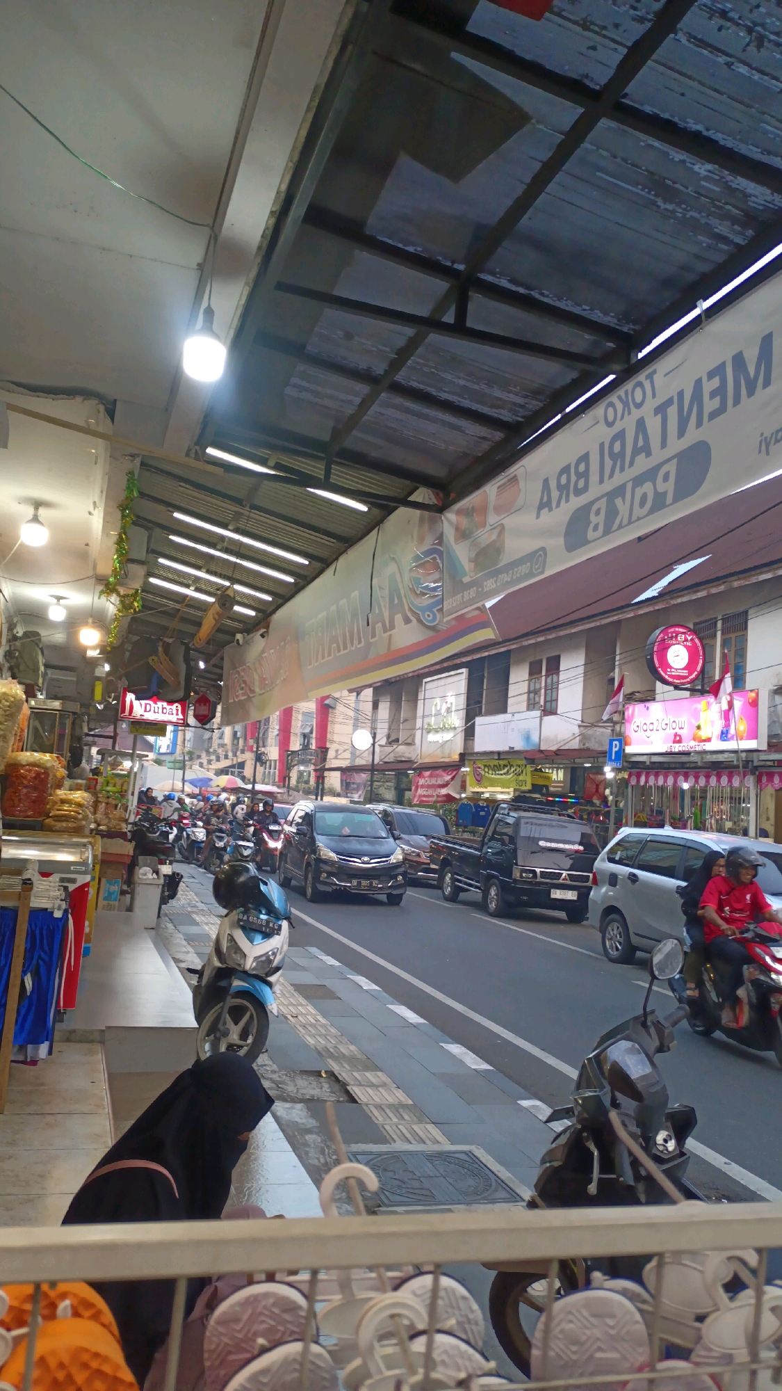 This image depicts a street scene in Padang Panjang, Indonesia, likely a commercial area. In the foreground, a woman wearing a black niqab and hijab is partially visible, seated near a sidewalk. Behind her, a white and blue scooter is parked. The street itself is bustling with traffic, including a black van, a black pickup truck, and a silver SUV, all moving forward. A person on a red motorcycle is also in motion.
The sidewalks are lined with shops, some displaying goods like clothing and snacks. A large banner stretches across the top of the scene, advertising a store with text that appears to be in Indonesian. Several streetlights and signs are visible, indicating an urban environment. The buildings along the street have multiple stories and are of varying architectural styles. The overall impression is one of a lively, active marketplace in a Southeast Asian city.