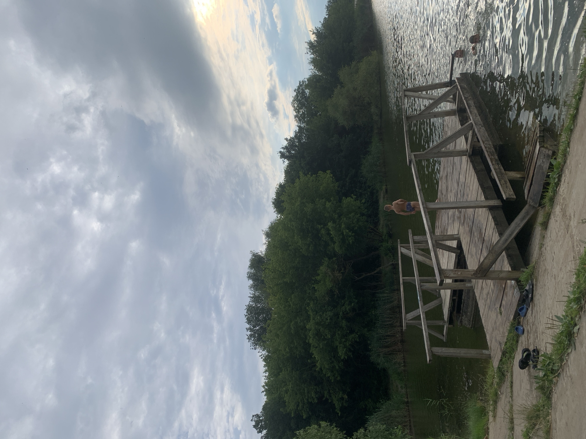 The image shows a wooden pier extending into a lake surrounded by dense green trees. The sky is cloudy with patches of sunlight. On the pier, a young boy stands facing the water. In the lake, two individuals are swimming. Several pairs of shoes and flip-flops are placed on the concrete ground near the start of the pier. The location is identified as a natural recreational area in or near Dondușeni, Moldova.