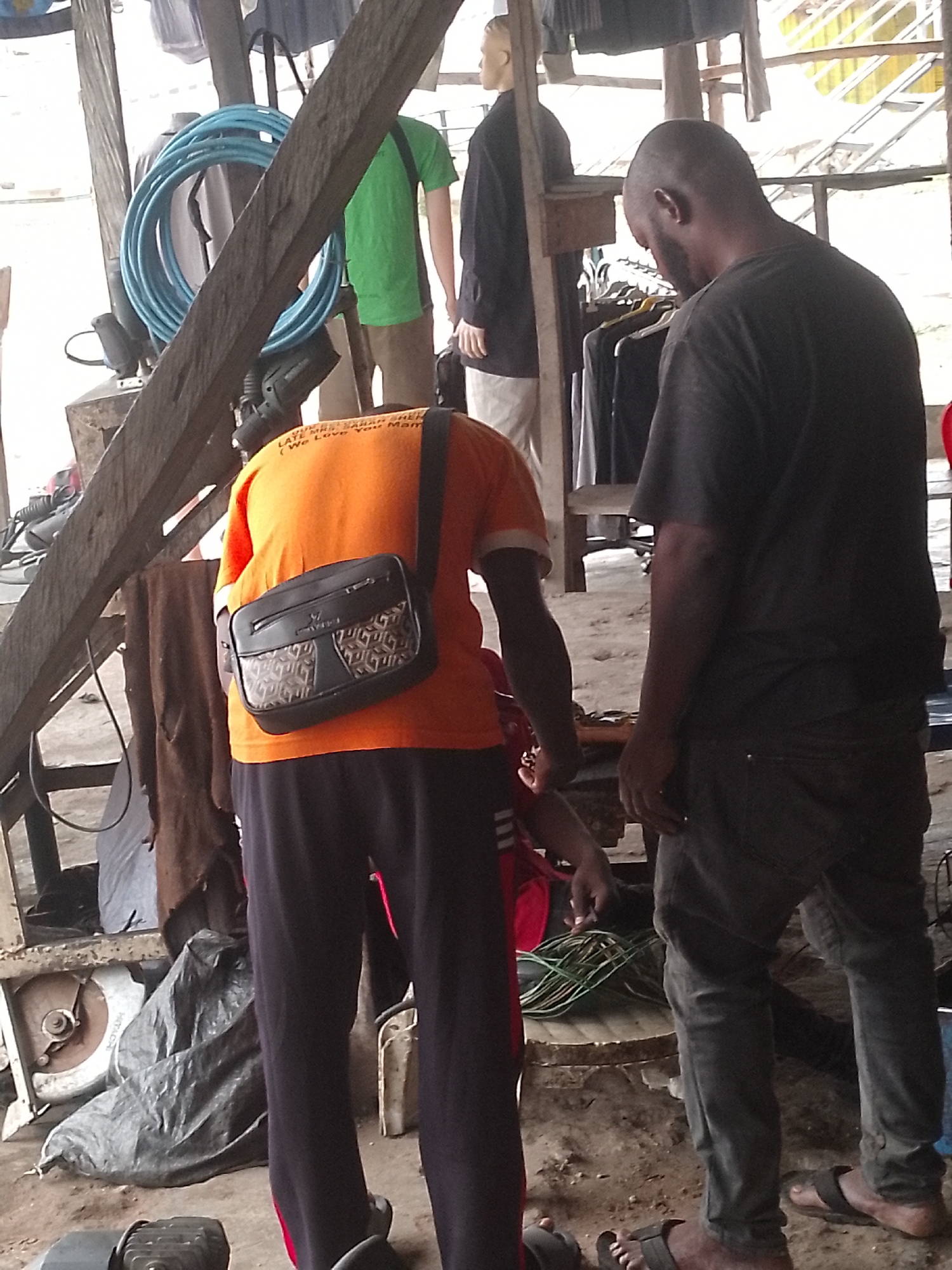 The image shows an outdoor scene in what appears to be a marketplace or informal business area in Lokoja, Nigeria. Two men are present. One is wearing an orange t-shirt with text on the back and carrying a black shoulder bag, and the other is wearing a dark t-shirt and jeans and looks down at the activity near his feet. There are mannequins wearing clothes in the background. Coiled blue hoses and power tools are visible, along with a table saw covered by a plastic bag. Wires are on the ground. The environment appears cluttered and functional.