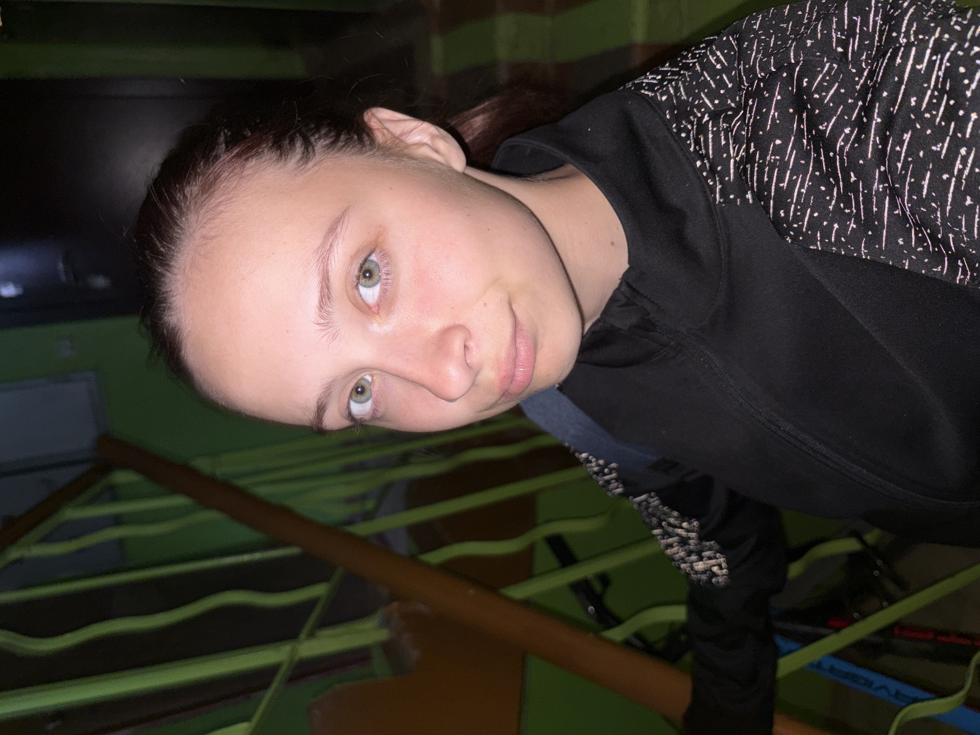 This image depicts a young female subject situated in what appears to be an indoor stairwell located in the city of Chaikovsky, Russia. The female has fair skin, dark brown hair pulled back, and distinct green eyes directed towards the camera. Her eyebrows are natural and light. Her lips are closed, presenting a subtle, slight smirk. She is wearing a black, long-sleeved athletic jacket with a zippered front closure. The sleeves and shoulder areas of the jacket feature a textured or patterned fabric composed of small white dashes and dots against a dark background. A dark blue strap, consistent with a backpack, is visible over her left shoulder. The background elements include a dark green wall and a stair railing system. A prominent brown wooden handrail extends diagonally across the frame, with vertical green bars visible behind it. A dark, rectangular panel or door is present in the upper right quadrant of the image. A partial blue object, resembling a bicycle frame, is visible in the lower left. The scene is illuminated by artificial, somewhat dim lighting.