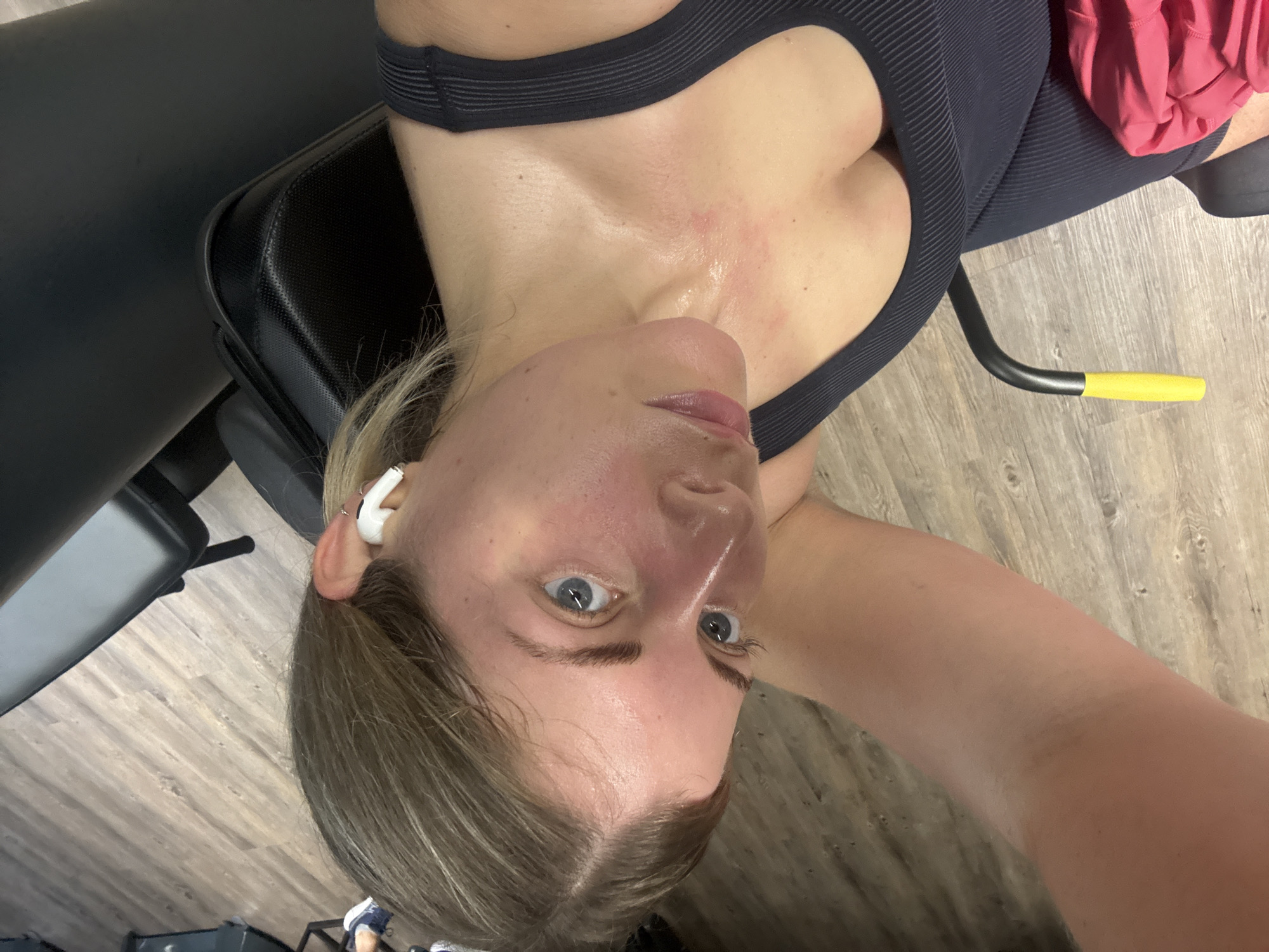 A high-angle, slightly rotated image captures a woman lying on her back on black gym equipment, likely taking a break during or after a workout in a gym in Paris, France. Her face, flushed and glistening with sweat, is central to the frame, with her blue eyes looking towards the camera. She has light brown hair, some strands damp with sweat, and is wearing white wireless earbuds in her ears.
Visible on her chest and neck are patches of redness and more sweat, indicating recent physical exertion. She is dressed in a dark, ribbed sports top or bra. To her right, a partial view of a pink garment, possibly shorts or a towel, is visible. The gym environment is suggested by the black cushioned equipment she rests on, and a black and yellow metal handle from another piece of machinery extends into the frame. The floor beneath her is made of light-colored wooden planks. The overall impression is one of post-exercise recovery or a brief pause in a strenuous activity. No text is visible in the image.