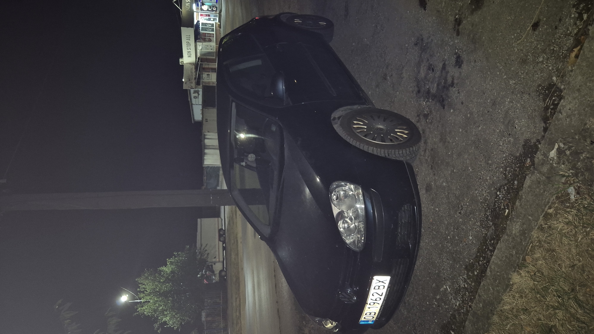 A dark-colored Volkswagen Golf hatchback is parked on a roadside at night. The car's license plate reads "OB 1962 BX". In the background, a lit building with a sign that says "NON STOP ALL" is visible, suggesting a commercial establishment. A streetlamp is also illuminated to the left of the car, casting light on a nearby tree and a small trailer. The ground is paved with asphalt and gravel, with some dry grass and a concrete curb. The scene appears to be in Lovech, Bulgaria. There are no people directly visible in the image, but a reflection in the car's windshield suggests there may be a person inside.