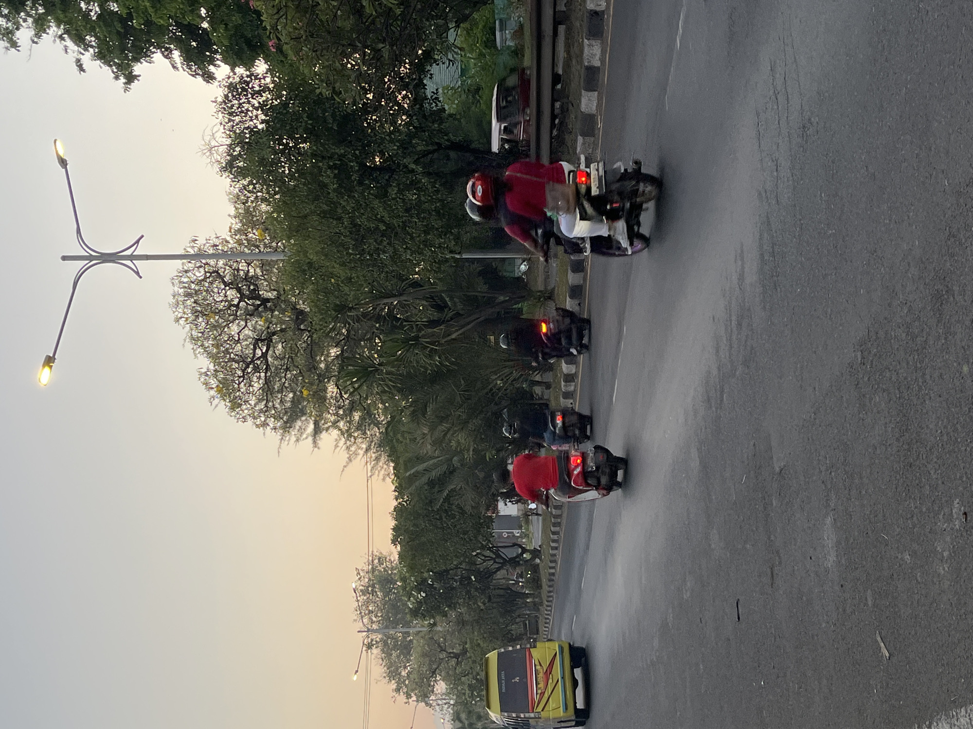 A photograph taken from a street-level perspective captures a road scene in Boro, Indonesia during the golden hour. The sky exhibits a gradient of pale yellow to light blue, suggesting either sunrise or sunset. A street lamp with two lights is prominently positioned in the upper right quadrant of the frame. The road itself is paved and appears wet, reflecting the ambient light.
Several vehicles are present on the road. On the left, a yellow minibus with "PAHAM JAYA" written on its rear is traveling. Further down the road, three motorbikes are visible. The first, a red motorbike, has a rider wearing a red shirt. Behind it, a motorbike carries two individuals, both wearing helmets, the passenger in a red shirt and the driver in a dark top. Further still, another motorbike with a rider wearing dark clothing is observed.
Lush green trees line the side of the road, with some trees bearing yellow flowers. A metal barrier separates the road from a slightly elevated area beyond. In the background, a red van can be seen parked. The overall impression is one of urban traffic in a tropical or subtropical environment.
