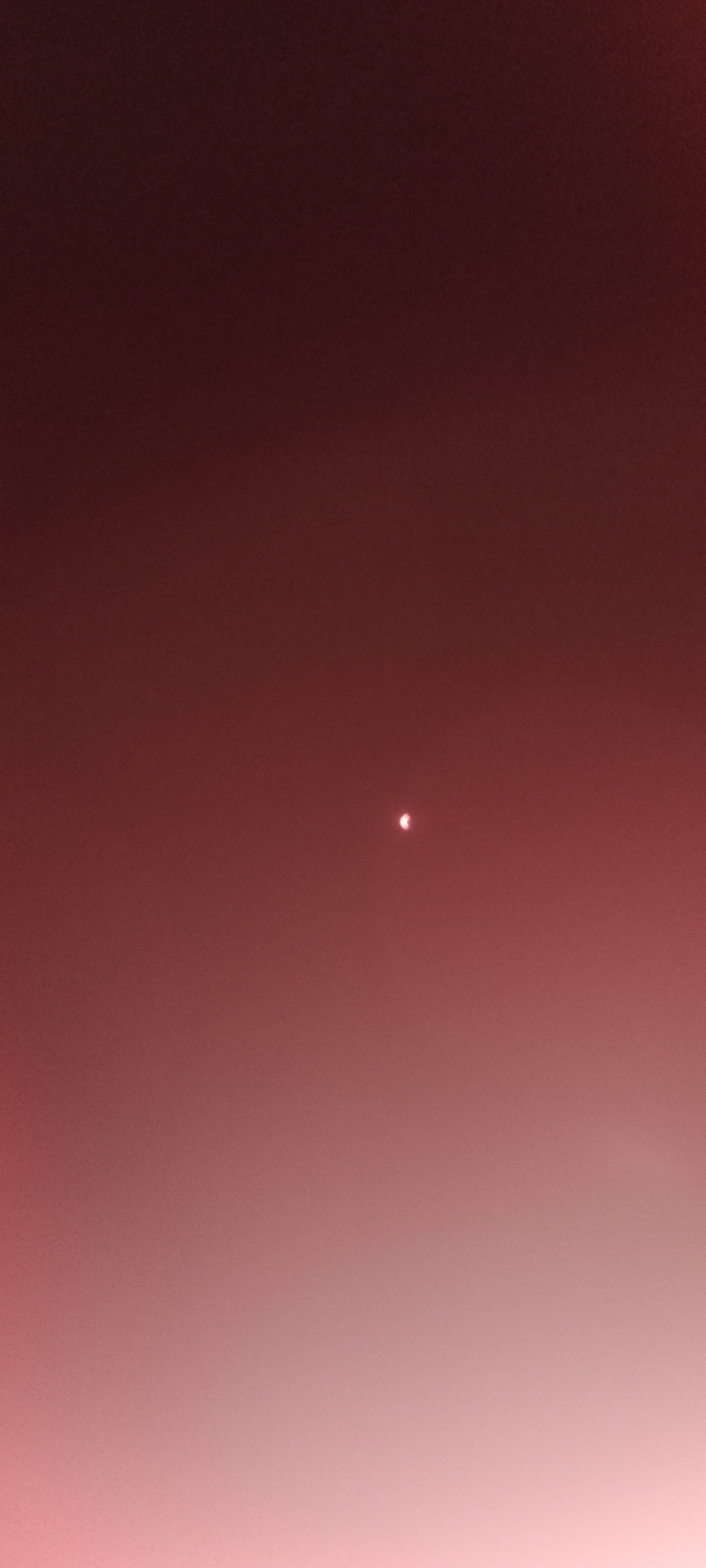 The image depicts a night sky with a diffused red or pinkish hue, possibly due to light pollution or atmospheric conditions. In the center of the frame, a small, bright celestial object is visible, appearing as a crescent or a partial sphere. The object is luminous and stands out against the darker background. The rest of the sky is largely uniform, with a gradient of color, becoming lighter towards the bottom of the image. There is no visible text, people, or specific identifiable objects or structures that would indicate a particular location or time of day beyond it being dark. The scene is purely celestial, focusing on the single bright object in the vastness of the sky. The overall impression is one of looking up at the night sky, with a solitary and notable celestial body. Given the provided location context of Adai, Spain, and the appearance of a celestial object, this could be a picture of a planet, a very bright star, or perhaps an artifact of a solar or lunar eclipse being photographed from that region.