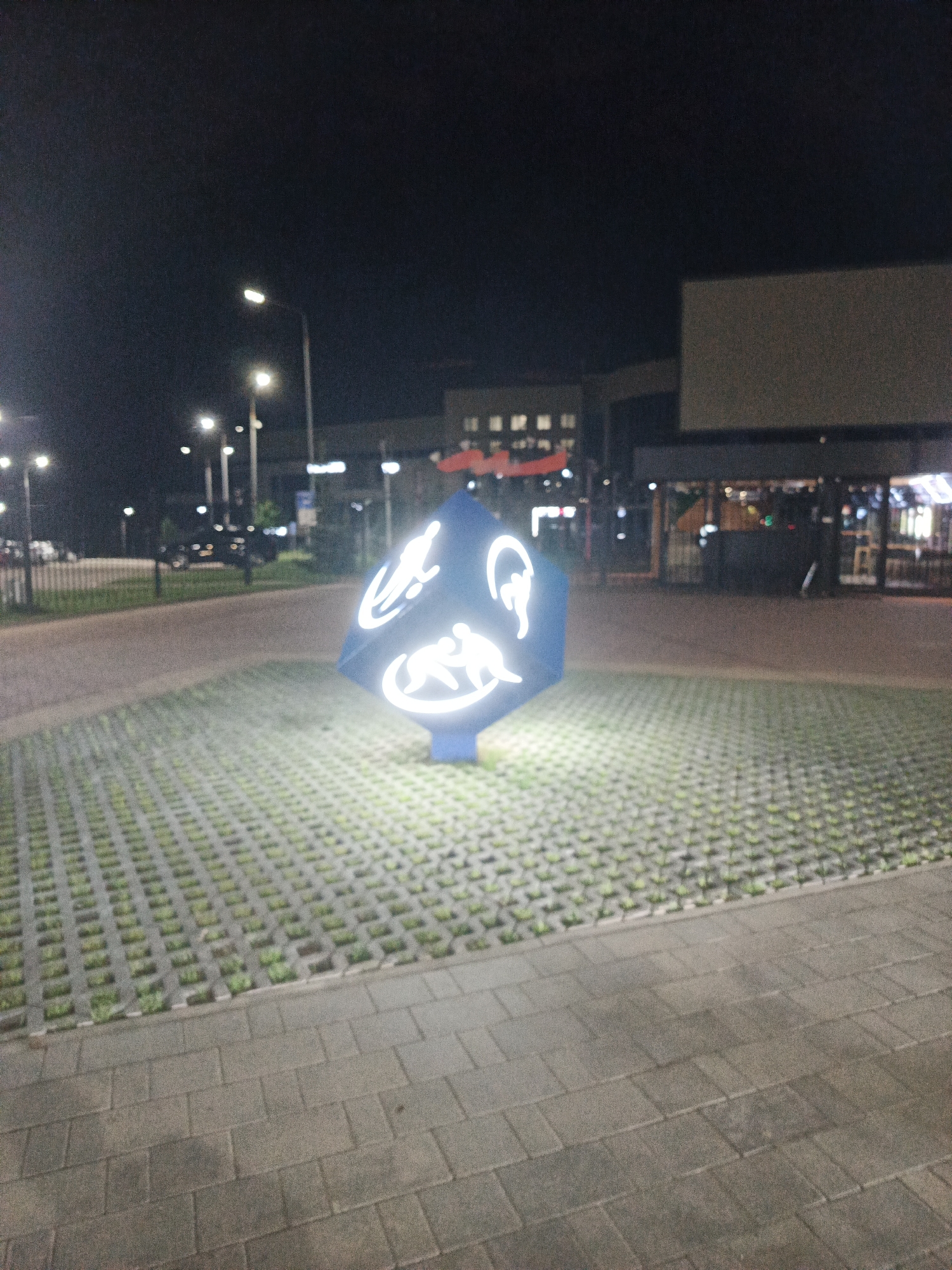 The image depicts an outdoor scene at night. The foreground features a paved area with interlocking concrete blocks, some of which have grass growing between them. To the right, a large, blue, angular sculpture is illuminated by bright white lights. The sculpture has white line art designs on its surfaces, resembling figures in motion. Behind the sculpture, a pathway or road is visible, lined with streetlights that cast a yellow glow. In the background, there are buildings with illuminated windows and signs, suggesting a commercial or urban area. The overall impression is that of a public space, possibly a parking lot or plaza, with decorative lighting.