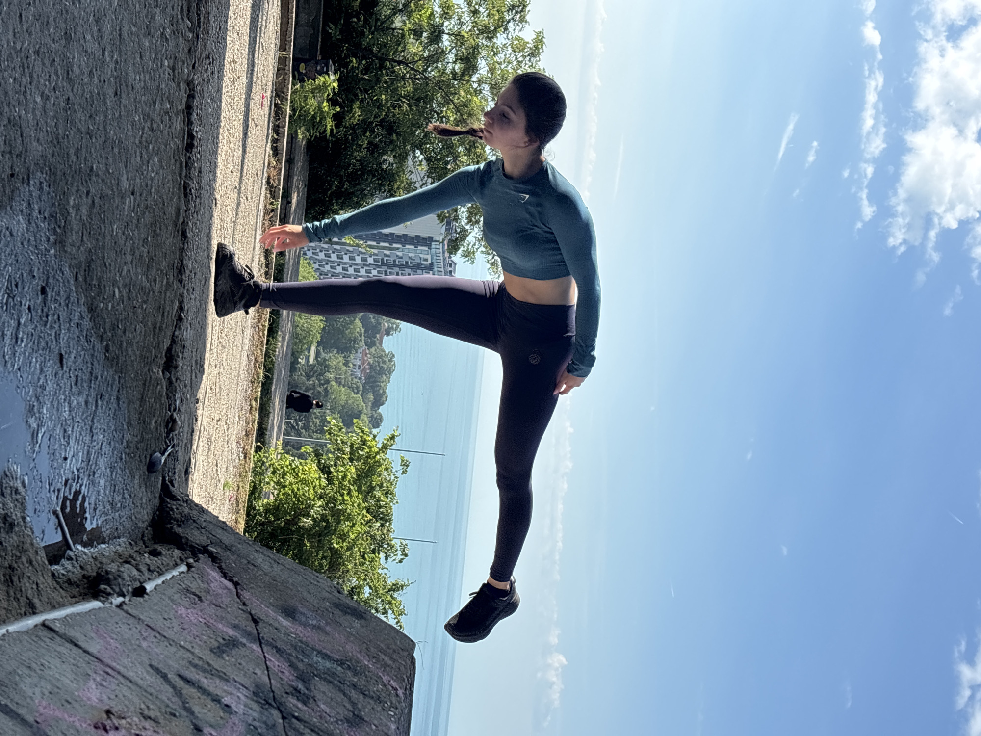 A young woman is shown performing a balance or stretch pose on a concrete ledge, overlooking a scenic coastal view. She is facing towards the right side of the frame, with her left leg extended high and her arms slightly outstretched for balance. Her dark hair is pulled back in a ponytail. She is wearing a long-sleeved teal cropped athletic top and dark navy athletic leggings, paired with black athletic shoes. A small white logo is visible on the left chest of her top, and another on her left hip on the leggings, characteristic of athletic apparel.
The scene is set outdoors under a bright, clear blue sky with scattered white clouds, indicating sunny weather during the day. In the background, a vast expanse of calm blue sea stretches to the horizon. Along the coastline, lush green trees and modern buildings are visible. The concrete surface in the foreground appears rugged and weathered, with visible cracks and some faint, indecipherable graffiti. The overall impression is that of a scenic, elevated viewpoint in a coastal city like Varna, Bulgaria.