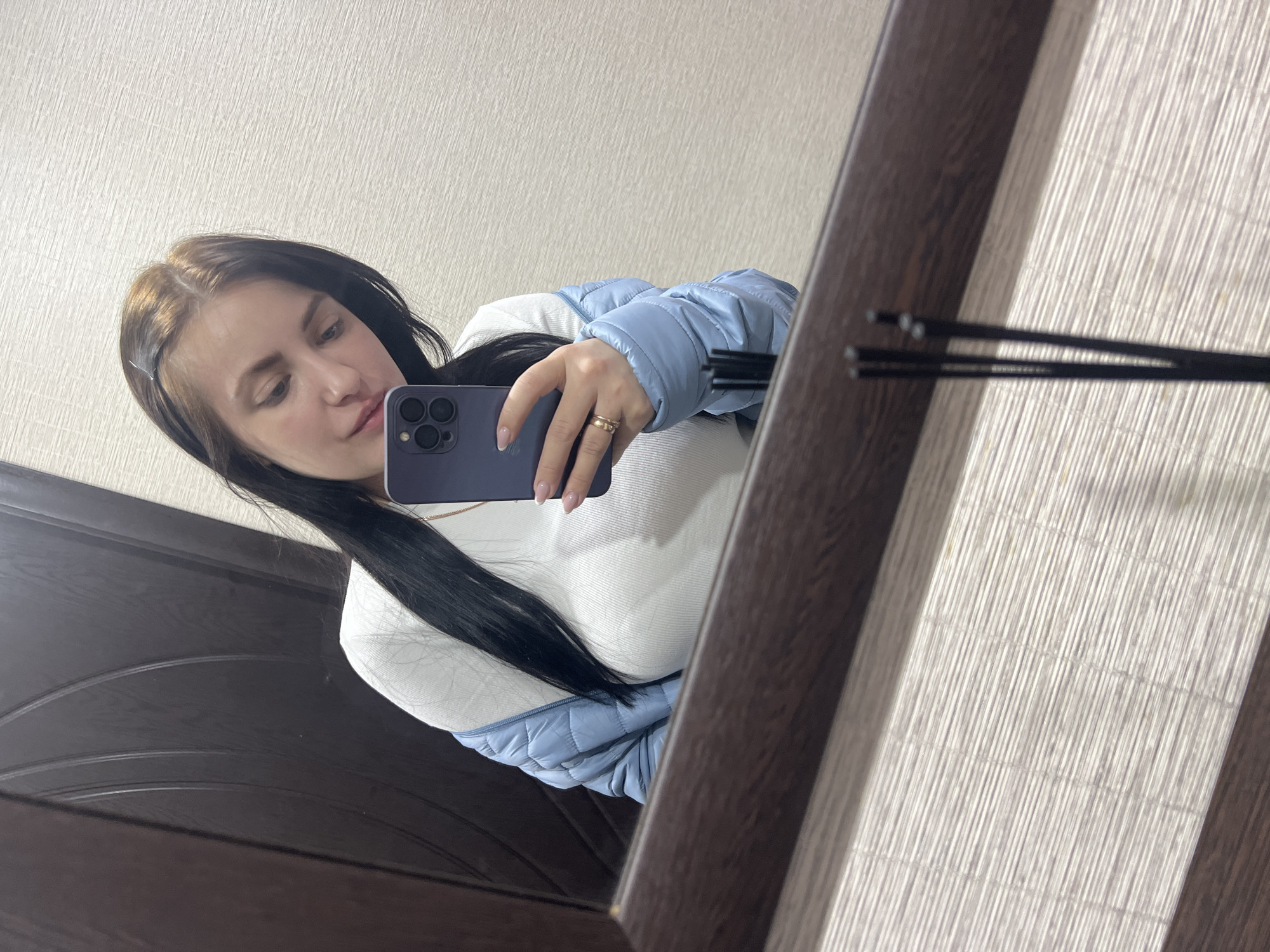 A woman is taking a selfie in a mirror. She has dark hair with light brown roots and is wearing a light blue puffer jacket over a white sweater. She is holding a purple phone in her right hand. She has two rings on her ring finger. The mirror frame is dark brown. The background includes a light-colored wall and a dark brown door. The location is Noviy, Russia.