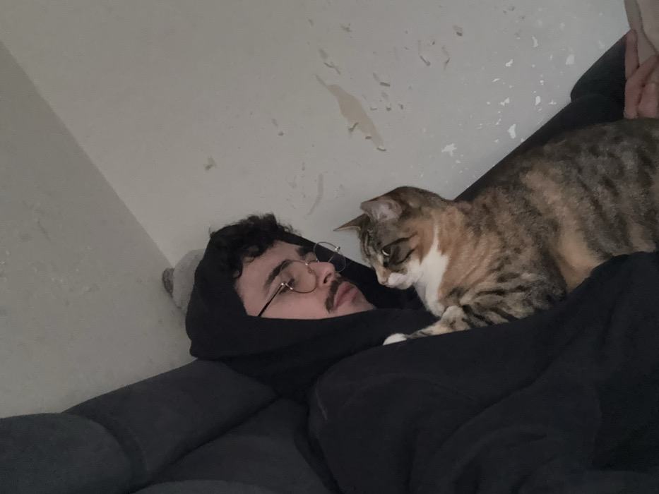 An indoor scene within a domestic residence in Rennes, France, depicts one adult male and one cat. The male, with dark curly hair, a mustache, and wearing round eyeglasses, is dressed in a black hooded sweatshirt with the hood up. He is reclined against a dark gray upholstered surface, his head slightly tilted downward, with his gaze directed towards the cat. The cat, a tabby with brown, black, orange, and white fur, is positioned against the man's left side. One of the cat's front paws rests on the man's sweatshirt. The cat's head is turned to its right, looking upwards and away from the man. The background consists of a light-colored wall with multiple irregular patches of peeling paint or surface texture.