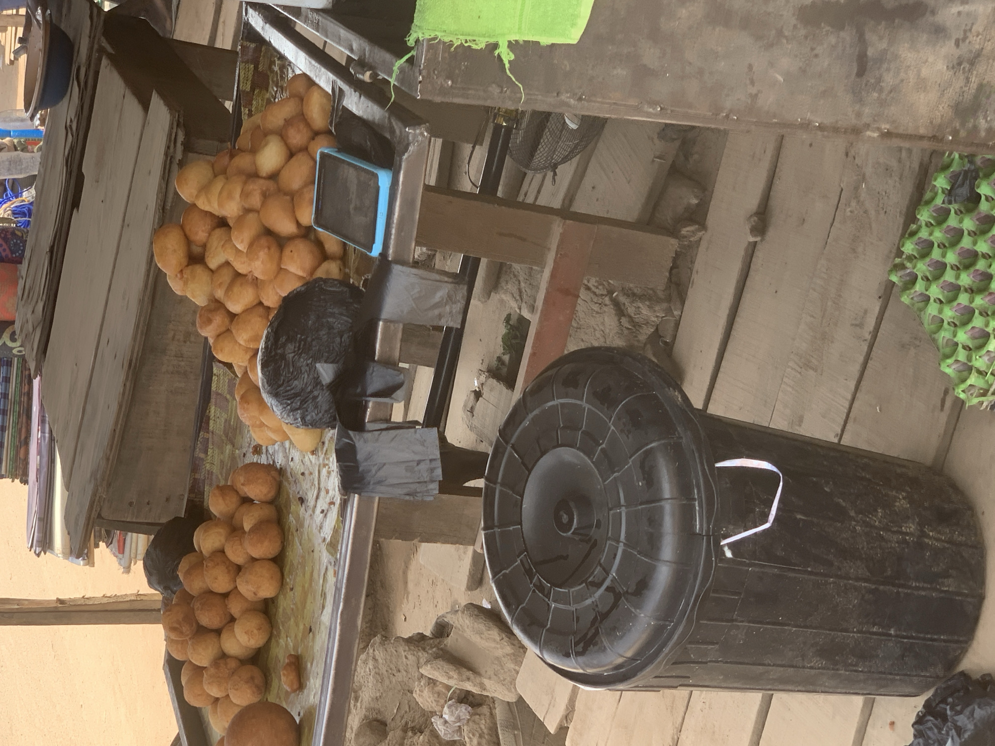 The image depicts an outdoor scene in Shagamu, Nigeria. A vendor's stall displays piles of golden-brown fried dough balls, potentially puff-puff. A small blue tablet sits among the food. Below the stand, a large black plastic trash bin lies on its side on the ground. The stall appears to be constructed from wood and metal. The ground is a mix of dirt, wood planks, and debris. A green cloth is partially visible above the stall, along with a small fan. Black plastic bags are nearby, possibly used for packaging.