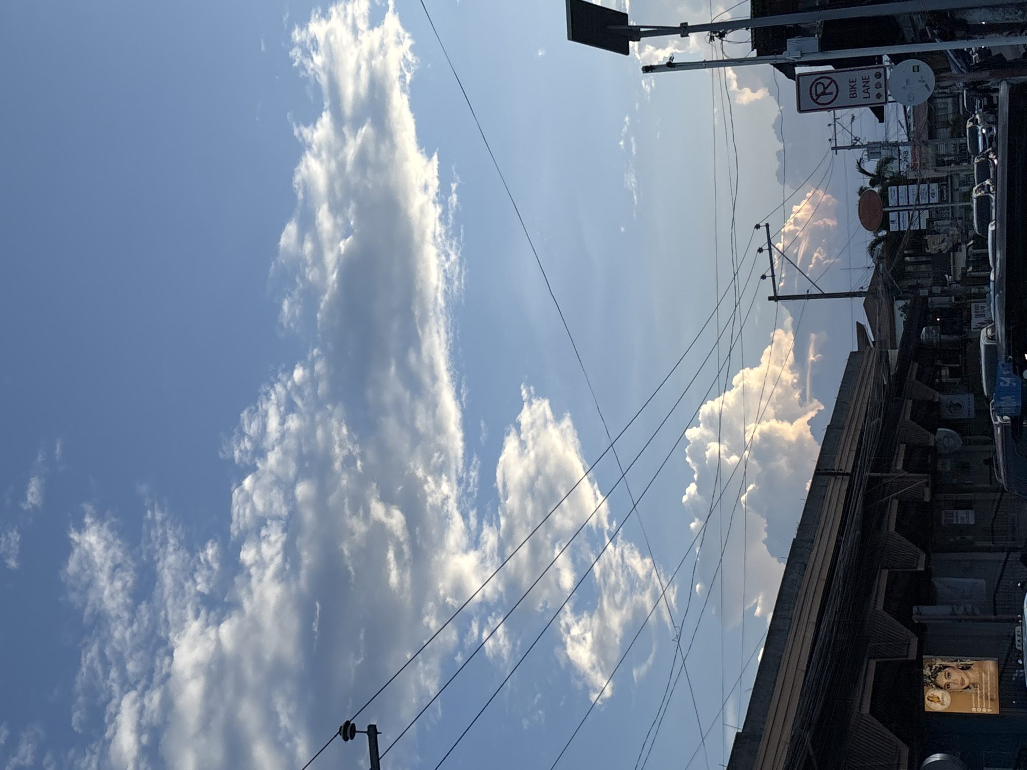 The image captures a street scene in Davao City, Philippines, under a partly cloudy sky. The foreground features a dark, commercial building with a sign displaying a woman's face and the word "LOVE." Power lines crisscross the sky, some attached to utility poles. A street sign indicating "BIKE LANE" and a "NO PARKING" symbol are visible on the right side of the frame. Vehicles, including a taxi, are present on the street. The background shows a cluster of buildings and trees, with large, puffy clouds dominating the sky, tinged with the warm colors of either sunrise or sunset.