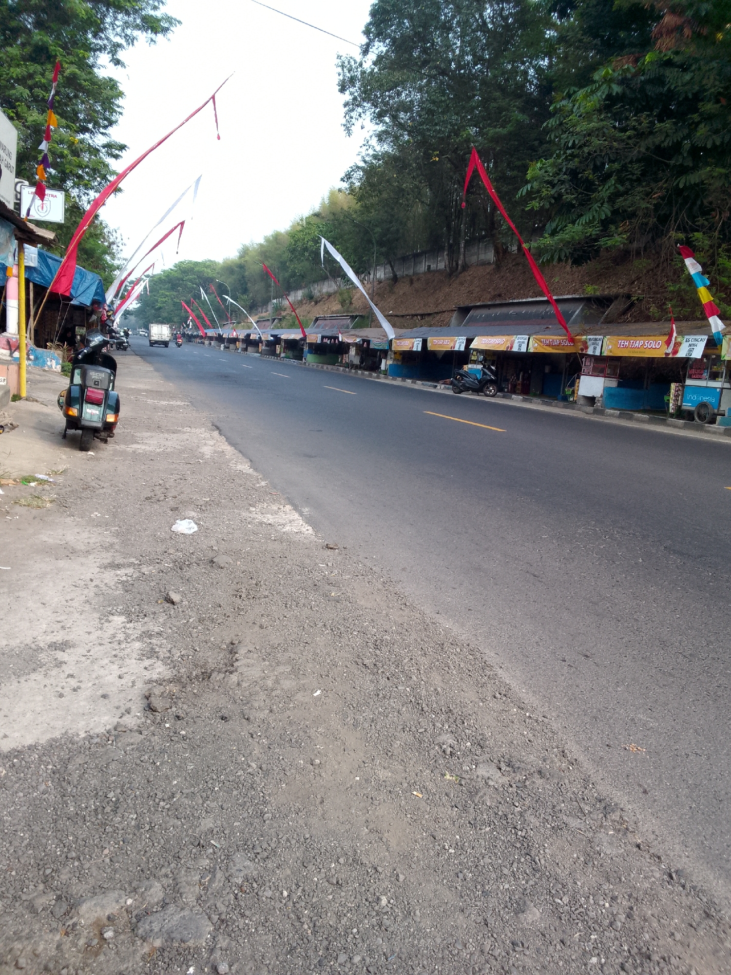 The image shows a street scene in Haurwangi, Indonesia, with several food stalls lining the road. Red and white flags, likely representing the Indonesian flag, are prominently displayed, suggesting a festive or patriotic occasion. A green scooter is parked on the side of the road, and a few people can be seen near the stalls. The road is asphalt, with a dusty and gravelly shoulder. Trees and lush vegetation can be seen on a hillside bordering the road. The stalls appear to be selling drinks and snacks, with signage indicating "TEH TIAP SOLO" (Solo Tea Every Day). A white cart with "Indonesia" written on it is also visible. The overall atmosphere seems to be that of a typical roadside market in a tropical environment.