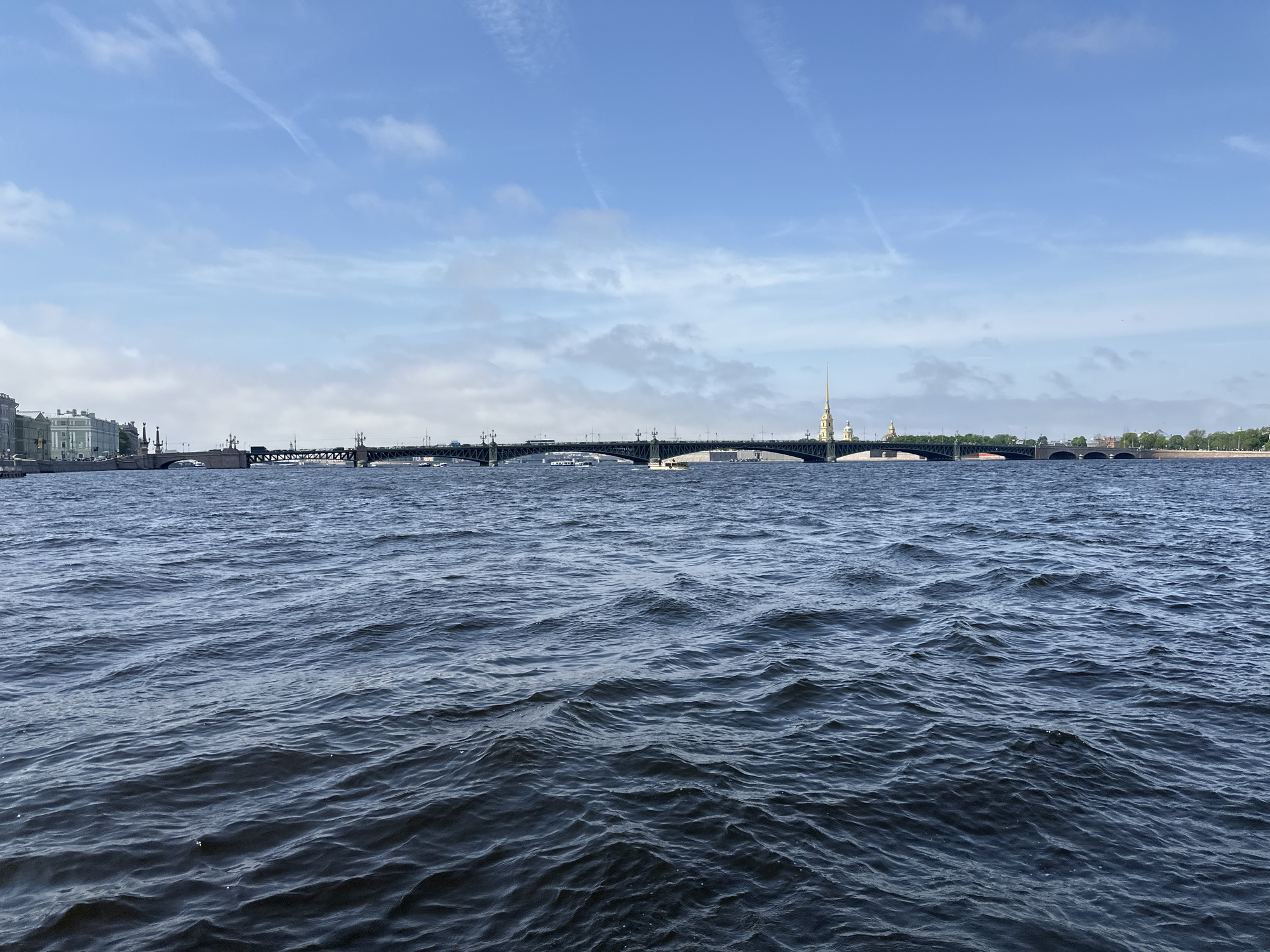 The image shows a body of water, likely the Neva River, in Saint Petersburg, Russia. A bridge spans the river in the middle ground, connecting parts of the city. In the background, the Peter and Paul Fortress, identifiable by its distinctive golden spire, is visible. The sky is blue with scattered clouds and contrails, suggesting a clear day. The water surface is rippled, indicating some wind or current.