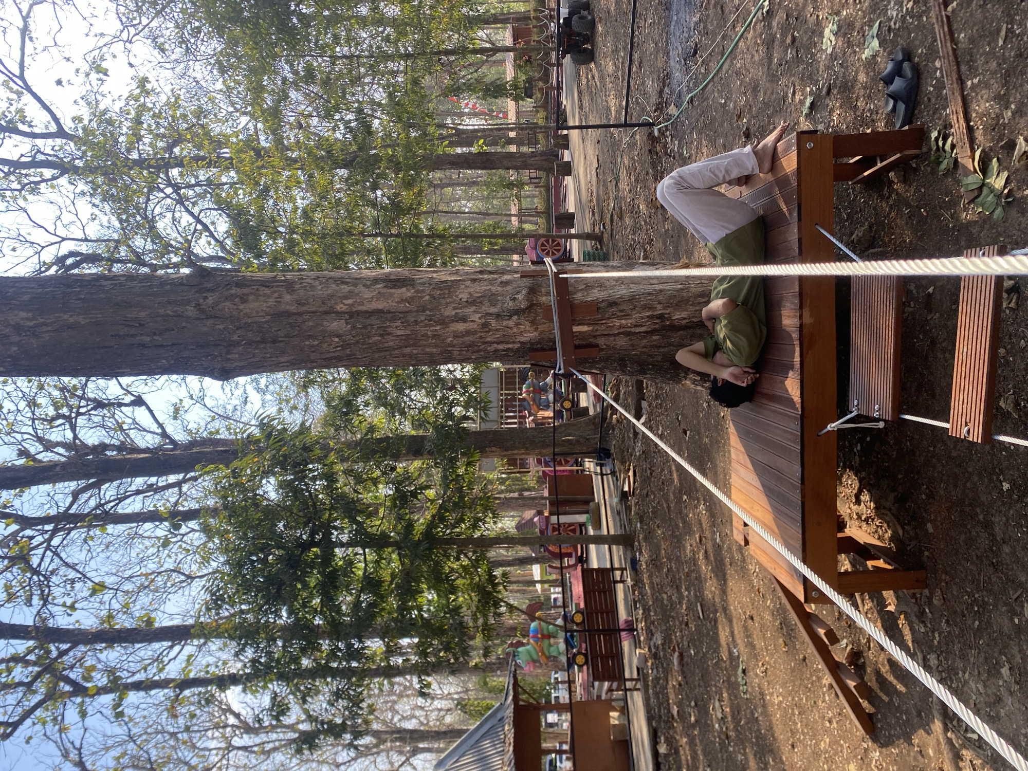 A person is lying on their back on a wooden platform in an outdoor area. The person is wearing a green shirt and light-colored pants. Their head is resting on their arm, and their eyes appear to be closed. The platform is supported by wooden legs and is situated near a large tree. A rope runs from the tree down to the platform and appears to be part of a rope ladder or bridge. In the background, there are more trees, some playground equipment with colorful figures, and a small building with a corrugated metal roof. The ground is covered in dirt and fallen leaves. The scene appears to be a park or recreational area. This is located in Ketileng, Indonesia.