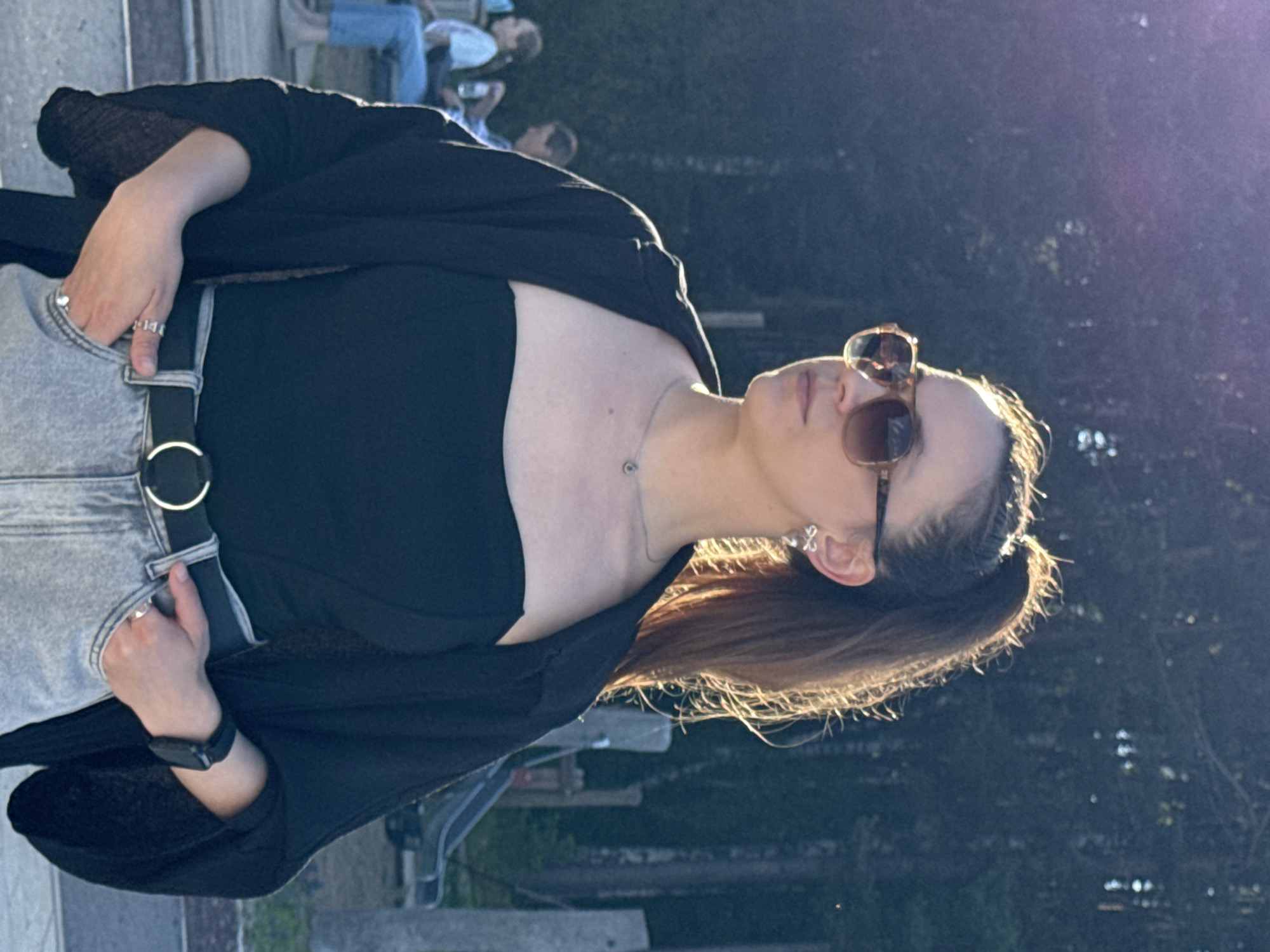 The image shows a young woman with her hair in a high ponytail, wearing sunglasses, a black strapless top, and a black open shirt. She has a silver necklace with a circular pendant and a pair of silver bow-shaped earrings. She is also wearing a black belt with a silver buckle and light-wash jeans. Her right hand is in her front pocket, and her left hand is also in her front pocket, with rings visible on her fingers. She is looking to her left. In the background, there are trees, some light posts, and two other individuals sitting on a bench, one wearing a white top and blue jeans, and the other a blue shirt. The scene appears to be outdoors, possibly in a park or recreational area. The lighting suggests it is late afternoon or early evening, with the sun casting a warm glow. The context of Chaikovsky, Russia is not visually discernible from the image itself.
