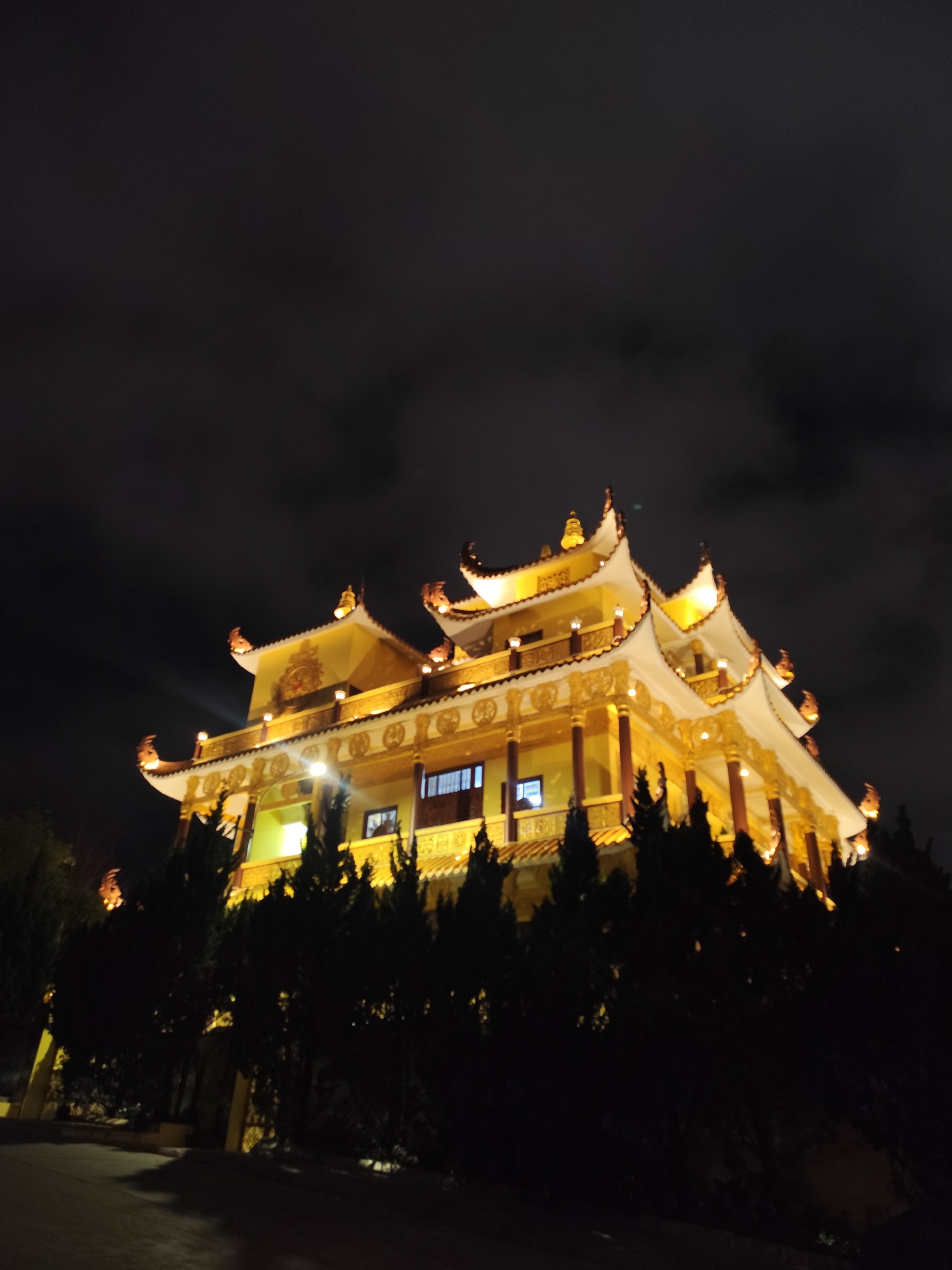 At night, a multi-tiered building illuminated with golden light dominates the view. The building has ornate, curved rooflines typical of East Asian architecture. It is partially obscured by a row of dark, coniferous trees. The sky above is black, hinting at nighttime conditions. The ground slopes gently and shows a paved surface. The location is most likely in Da Lat, Vietnam, given the architectural style. No people are visible in the image.