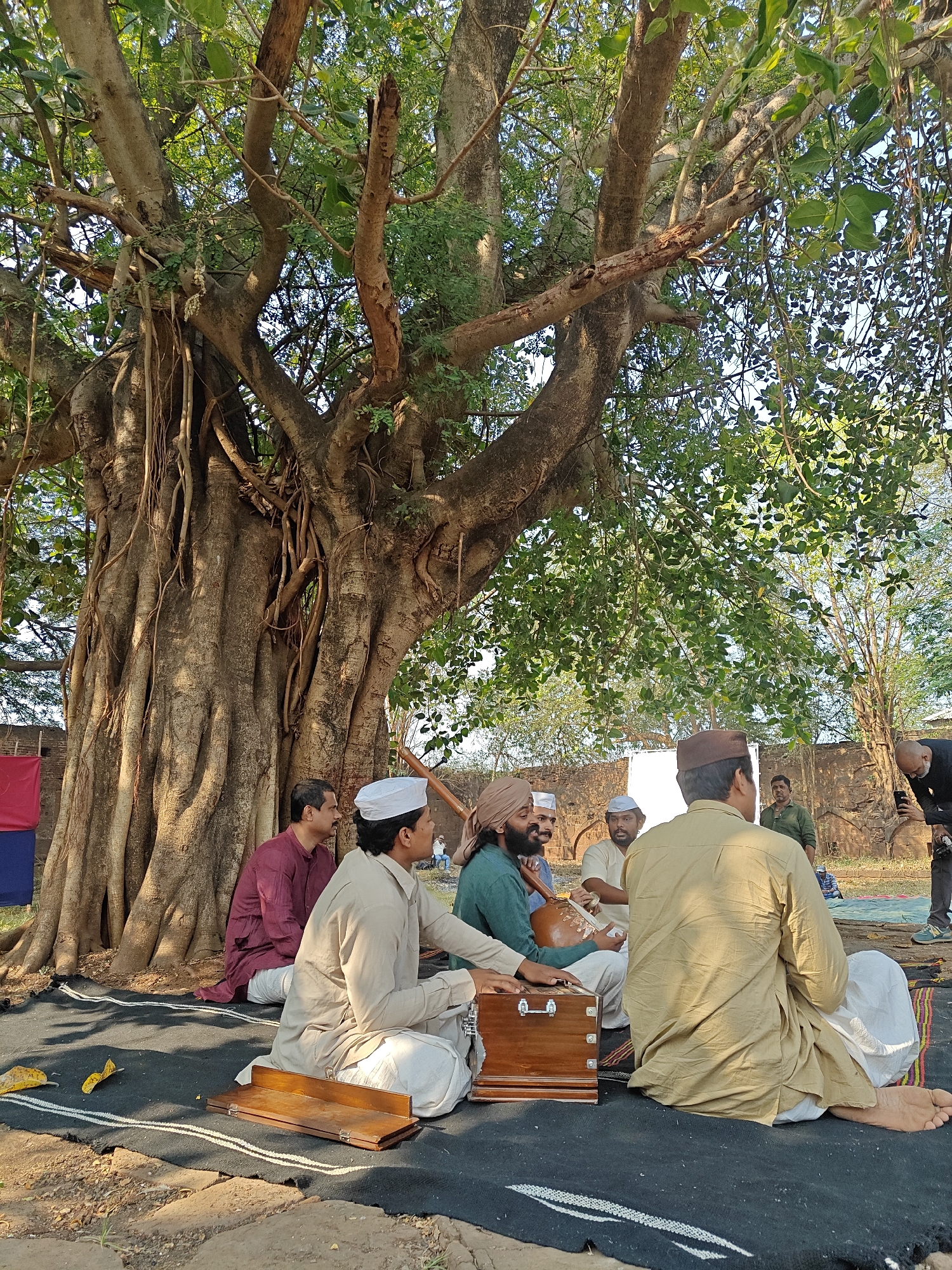 The image depicts an outdoor gathering of eight men under a large banyan tree at what appears to be a historical site or fort area within or near Kolhapur, India. In the background, ancient stone/brick walls with arched openings are visible.
Six men are seated on a black mat on the dirt ground in a semi-circle, facing towards the right side of the frame. One man in a light brown kurta and white cap, positioned center-left, is seated in front of a wooden harmonium. To his left, another man in a teal-green kurta and tan turban, with a beard, holds a gourd-shaped stringed instrument, likely a tanpura. A man in a maroon kurta sits behind them. A man in a light brown kurta and a white cap is seated further back, observing. In the right foreground, a man in a light brown kurta and brown cap is seated with his back to the viewer, his bare feet visible. A sixth man in a light green shirt is seated further back on the right.
The banyan tree dominates the left and upper parts of the image, featuring a massive trunk, aerial roots, and dense green foliage, providing shade. On the mat near the harmonium, a small wooden board and two yellow leaves are present. In the background, a man in a dark shirt is standing, bent over, holding a smartphone, seemingly capturing the scene. A white photography reflector disc is partially visible behind the seated group, along with a piece of red and blue fabric near the tree trunk on the far left. The scene is illuminated by natural daylight, casting soft shadows.