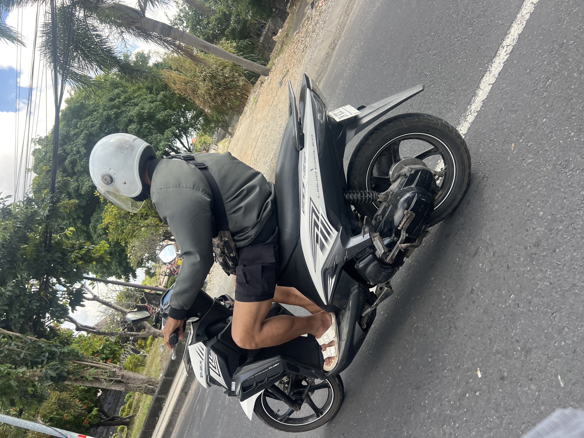 A man wearing a white helmet and a dark grey jacket is riding a black and white scooter on a paved road in Kuta, Indonesia. He is wearing black shorts and white sandals. The scooter has the "YAMAHA" logo visible on the front. The background shows lush green trees, palm trees, and some buildings. The sky is partly cloudy. The road has a white painted line.