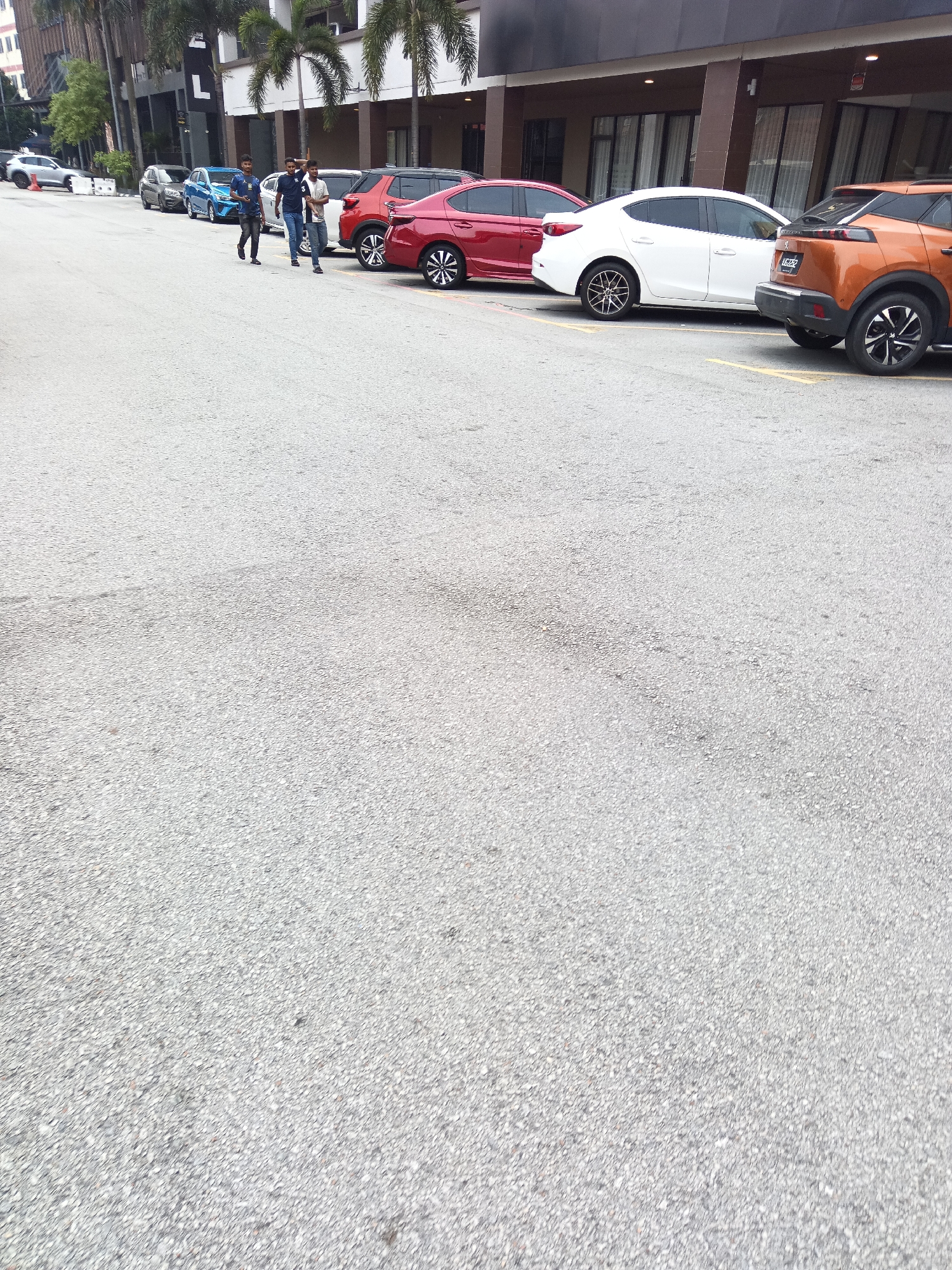 In a Kuala Lumpur, Malaysia cityscape, three men are walking on a paved road alongside parked cars. The men, of South Asian descent, are casually dressed. The first man on the left wears a dark t-shirt and dark pants. The second man in the middle wears a blue t-shirt and dark pants. The third man on the right wears a white t-shirt and dark pants. They appear to be walking from left to right, side by side, and are in conversation.
A row of cars is parked on the right side of the road. From front to back, the cars visible are a white sedan, a red sedan, and an orange SUV. The red sedan has a license plate that reads "VGG 7226". The parked cars are aligned with yellow parking lines on the asphalt.
Behind the cars, a building with dark-tinted windows and an awning is visible. Palm trees and other greenery are present in the background, suggesting a tropical urban environment. The road surface is asphalt with visible texture and some tire marks. The sky is overcast, indicating a potentially cloudy day.