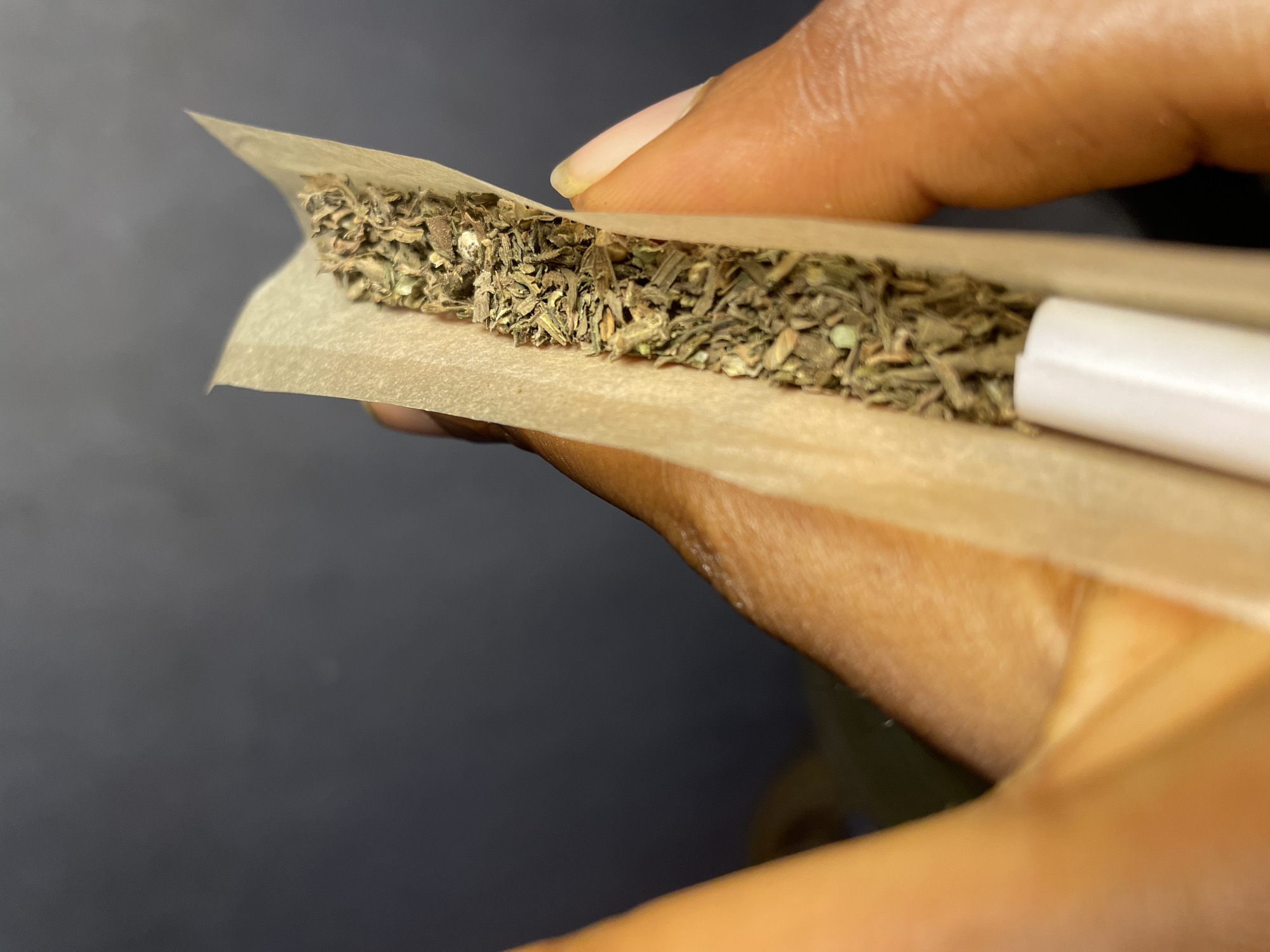 The image shows a close-up view of a person's hands rolling what appears to be a marijuana cigarette, also known as a joint. The hands are holding a light brown rolling paper filled with a green and brown plant material. A white filter or crutch is visible at one end of the paper. The background is dark and blurry. There is no information to suggest the location to be Nkpor, Nigeria.