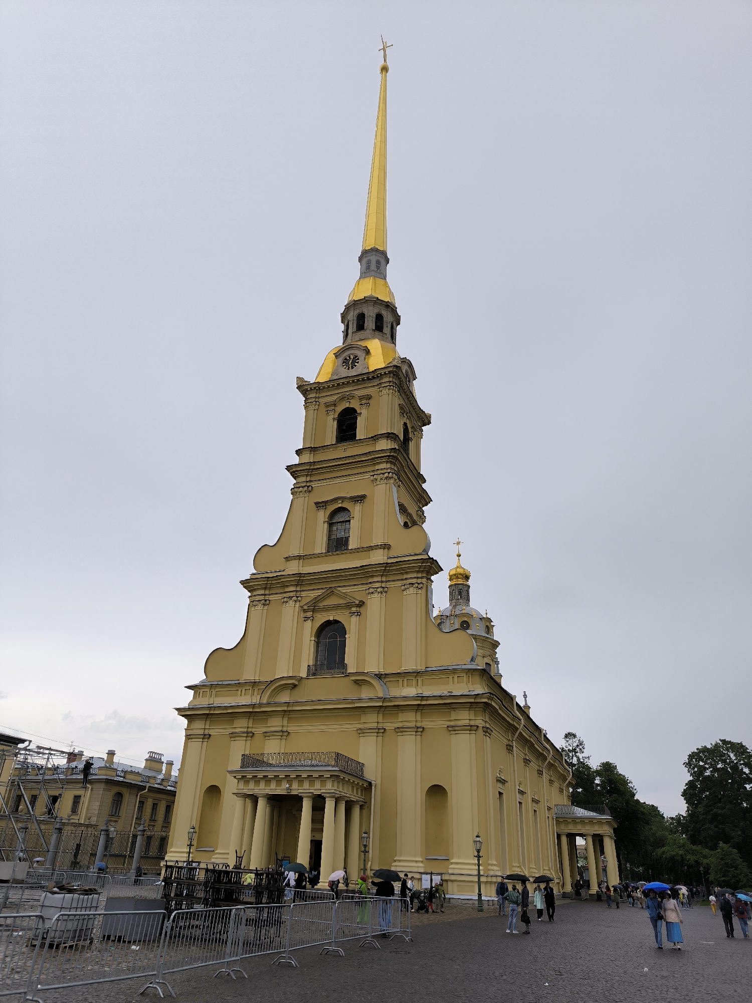 The image shows a tall, pale yellow building with a golden spire topped with a cross. The building has multiple levels and several arched windows. In front of the building, there are people walking, some holding umbrellas. There are also metal barriers and some construction equipment. The sky is overcast. The building is the Peter and Paul Cathedral, located in the Peter and Paul Fortress in Saint Petersburg, Russia.