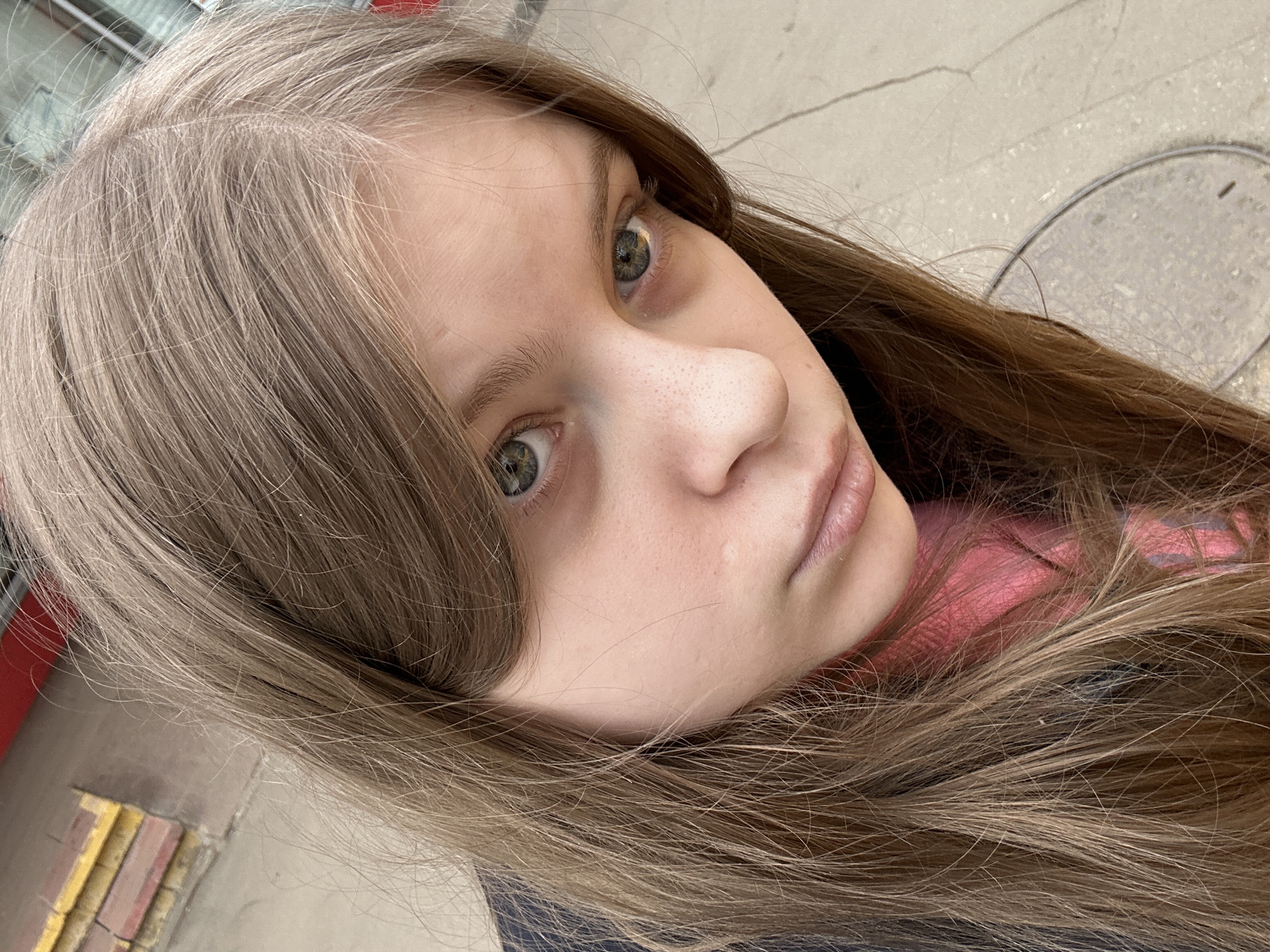 A close-up, upward-angled shot of a young Caucasian woman with light brown hair, looking directly at the camera with slightly upturned, greenish-gray eyes. Her complexion is fair, and a section of a pink garment is visible at the lower right. The background shows an urban setting with a portion of a sidewalk, including a metal manhole cover at the right edge. In the bottom left, the edge of what appears to be a train platform with yellow and red brick tiles is visible, likely in the city of Moscow, Russia.