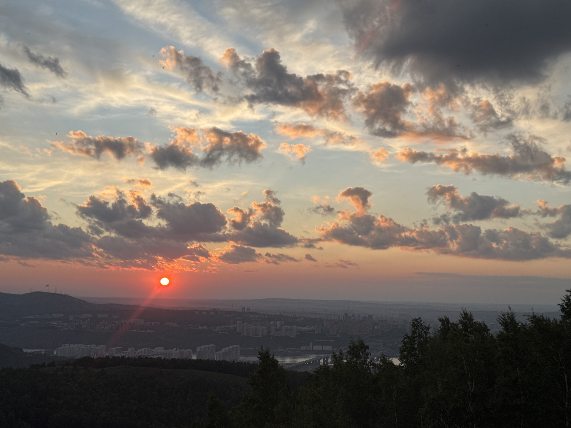 This is a scenic photograph of a sunset over the city of Krasnoyarsk, Russia, viewed from a high vantage point overlooking the city. The sun, a bright red orb, is low on the horizon, casting a warm glow and a sunbeam that extends downwards. The sky is filled with dramatic clouds, illuminated in shades of orange, pink, and grey, contrasting with the softer blue hues. In the middle ground, the urban landscape of Krasnoyarsk unfolds, featuring numerous buildings and a bridge spanning a body of water. The foreground is dominated by dark silhouettes of trees, framing the view of the city and the sky. The overall impression is one of a vast, natural spectacle occurring over a populated area, capturing the beauty of twilight.
