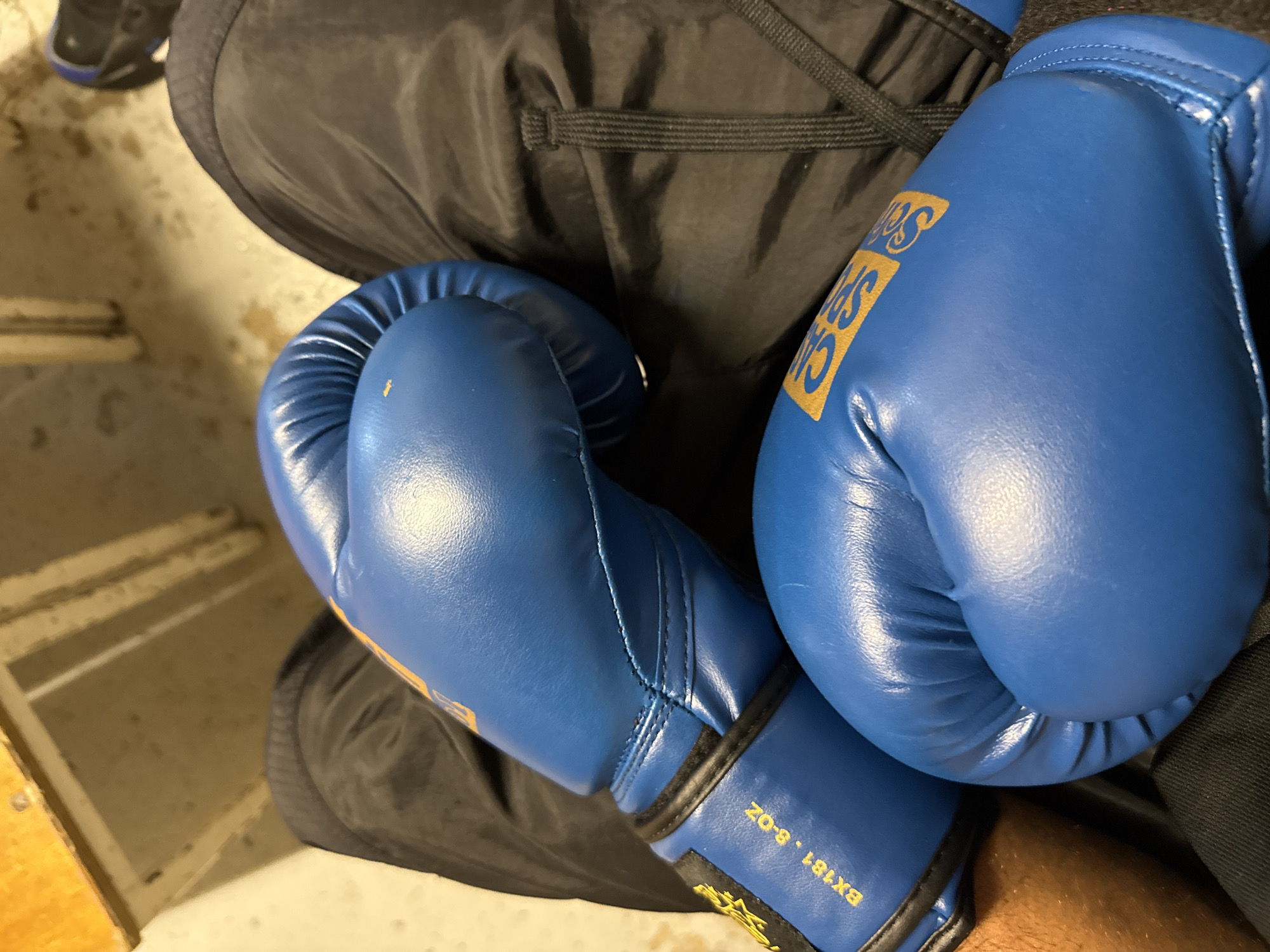 A close-up shot shows a pair of blue boxing gloves resting on a person's lap. The person is wearing dark-colored sweatpants with a drawstring waist. The gloves have gold lettering and a black trim around the wrist. The lighting suggests an indoor environment. No information is provided that indicates this image was taken in Sarcelles, France.