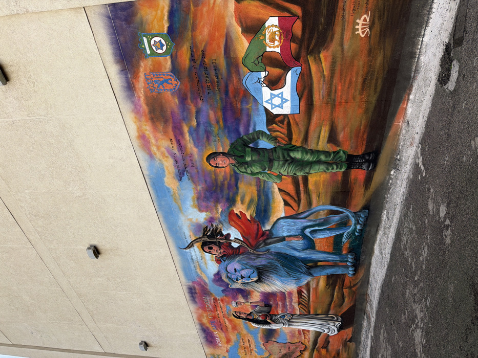 This forensic description details a large mural painted on the side of a light-beige building wall in Tel-Aviv, Israel, specifically near Dizengoff Square. The mural occupies the lower portion of the wall, with an unpainted, plain beige section above it. The foreground shows a dark, asphalt-paved surface.
The mural's background features a stylized desert landscape with undulating hills under a vibrant sky transitioning from blue at the top to hues of red, orange, and purple.
From left to right, the mural depicts:
* A woman in a long, flowing white garment, possibly traditional, with dark hair. She holds a white object (resembling a flower or dove) in her raised right hand and a red heart-shaped object in her left hand. To her right and slightly behind her, a light-colored outline of a geographical map is visible with some faint internal markings. Above this, the English text "WOMAN LIFE FREEDOM" is painted, accompanied by Hebrew text below it.
* A large, stylized blue lion with a prominent mane, facing right. A dark-haired figure, likely a woman, wearing a red tunic and a red cape, is depicted riding the lion. She holds a drawn bow with an arrow aimed upwards and to the left.
* To the right of the lion, a woman with dark hair, wearing a complete green military-style uniform and black boots, stands facing forward. Her hands are positioned at her waist, and a subtle golden halo-like glow surrounds her head.
* In the lower right, two hands are depicted forming a heart shape. The left hand is stylized with the design of the Israeli flag (blue Star of David on a white field), and the right hand features the design of the Iranian flag (green, white, and red horizontal bands with the Emblem of Iran).
* Above the hands and to the right, Hebrew text is visible, followed by "Jeremiah 49:39." Further to the upper right corner, two shield-shaped emblems are painted: one orange with a blue lion rampant, and another green with a gold Star of David and other symbols.
* Additional Hebrew text is present in the upper left section of the mural, followed by "Isaiah 9:5."
* Artist signatures or social media handles, including "@Hooma_" and "@the_graffiti_," are visible in the lower right corner.